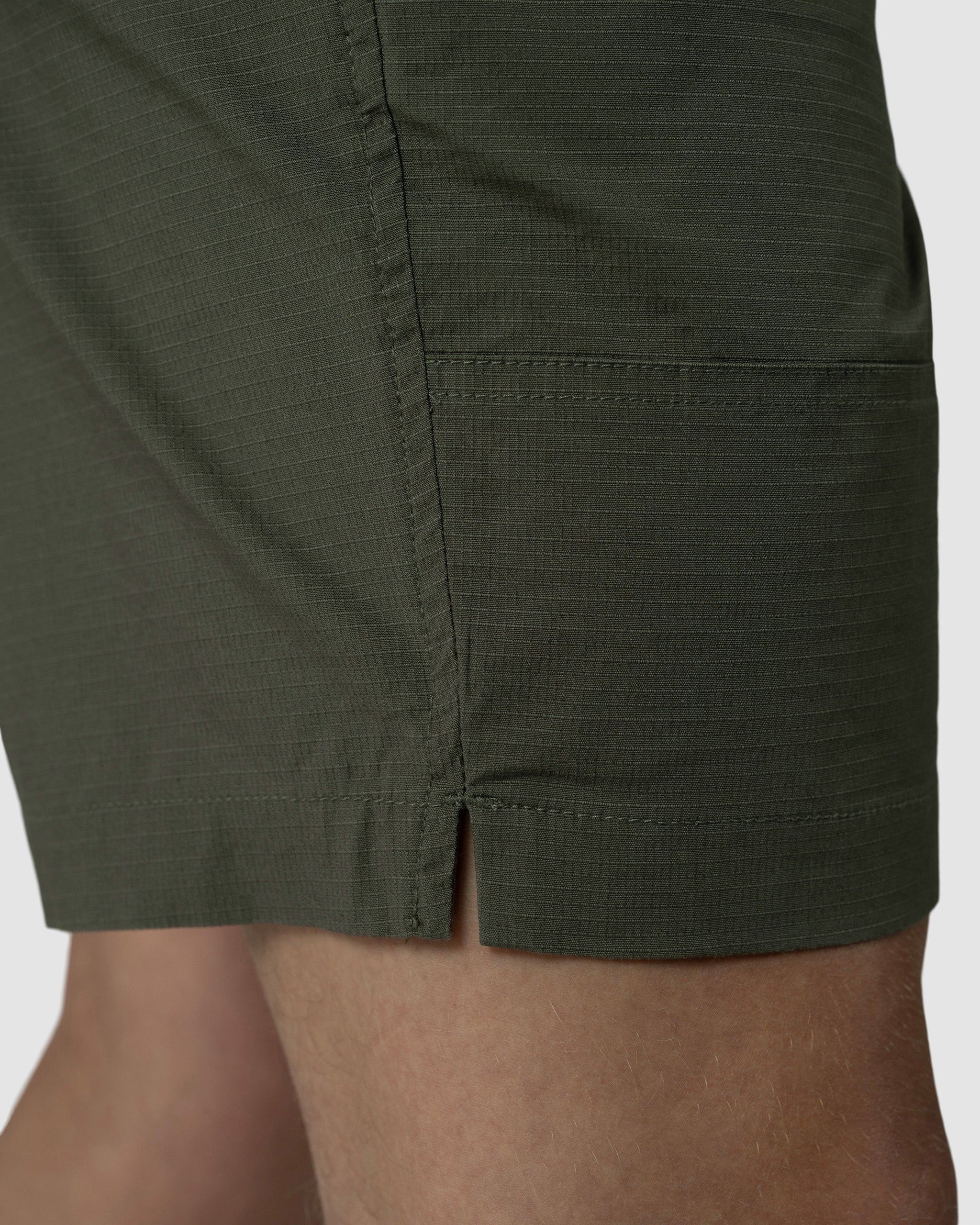Close-up of the lower side of a person wearing BEARDEDGOATs Mens Canopy Short in dark green stretch cotton ripstop, highlighting the small side slit and visible seam details against a plain background.