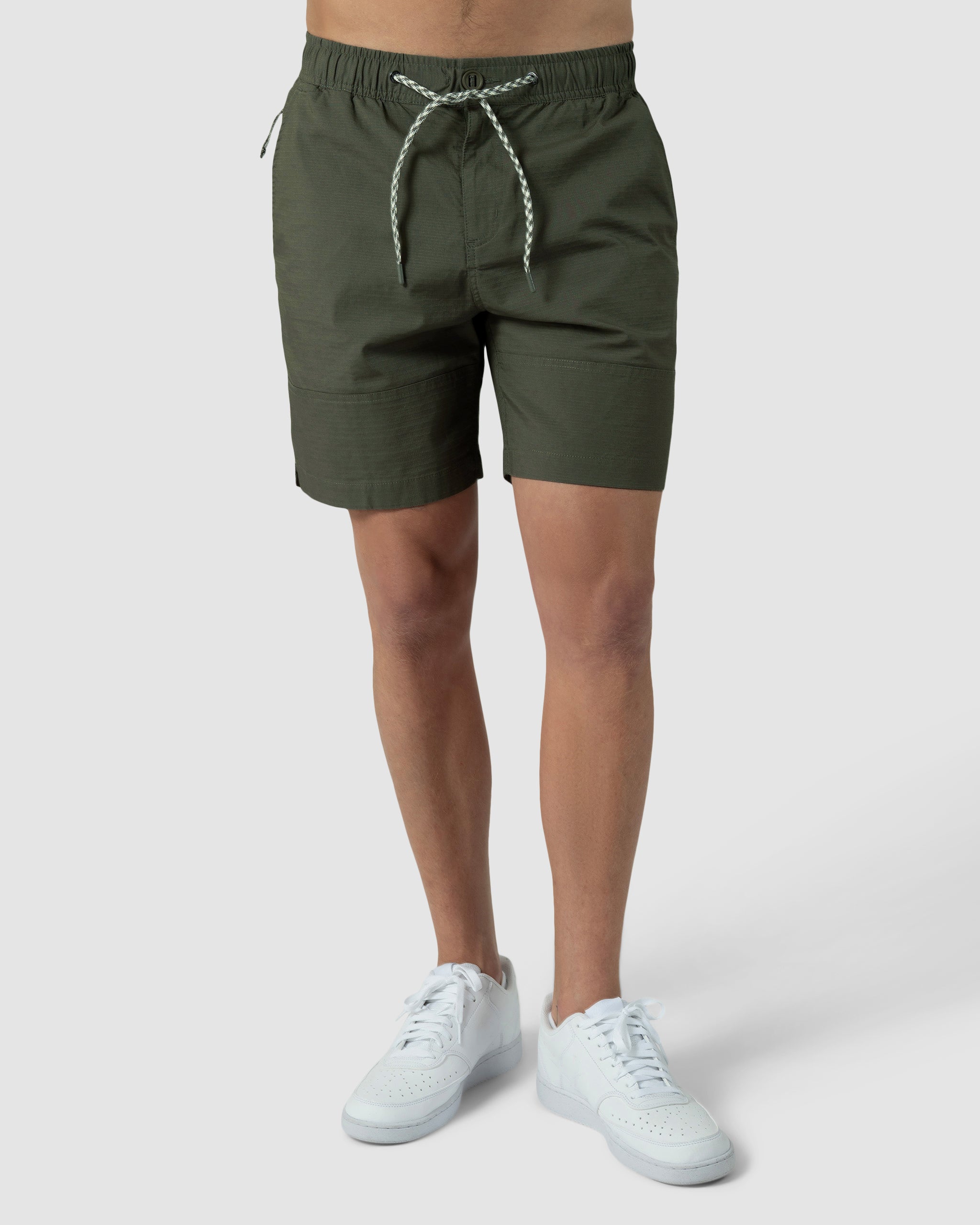 A person wears BEARDEDGOAT Mens Canopy Short in olive green, made from lightweight stretch cotton ripstop, with white sneakers. Only the lower torso and legs are shown against a plain light gray background.
