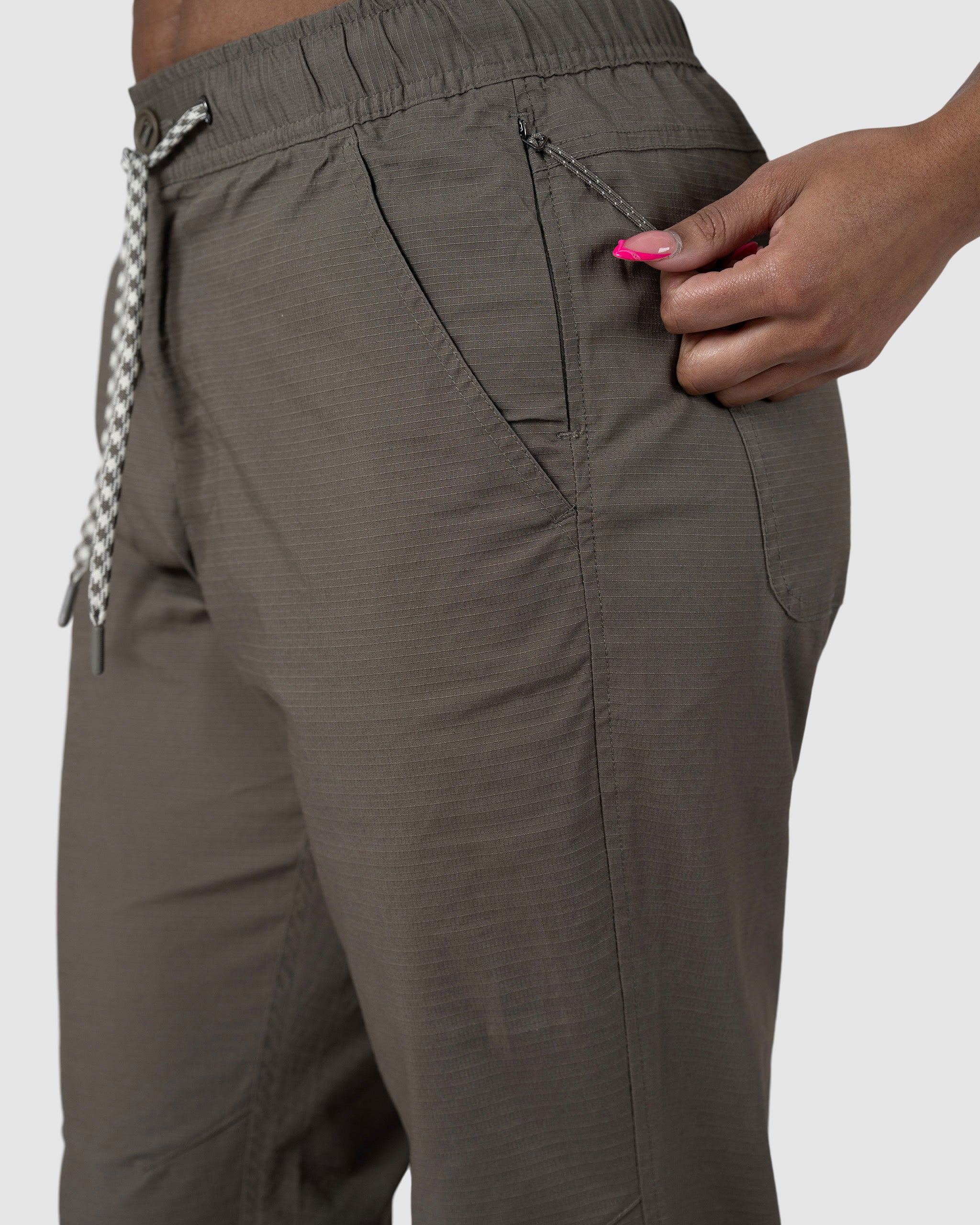 A person with pink nail polish unzips the side pocket of olive green BEARDEDGOAT Womens Canopy Pant drawstring pants, shown from waist to mid-thigh.