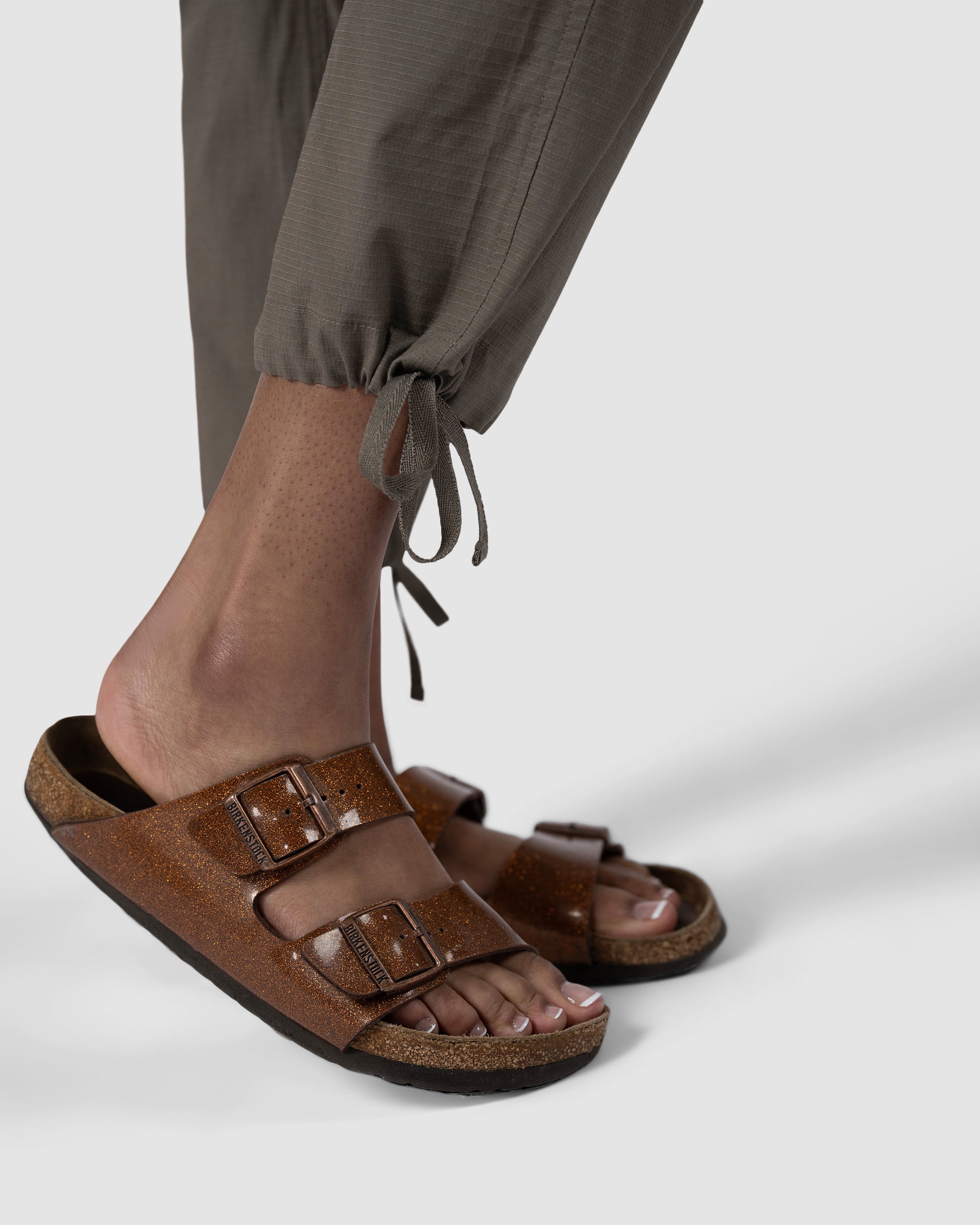 A close-up of a person wearing brown two-strap sandals and BEARDEDGOAT Womens Canopy Pant in olive green stretch cotton with adjustable ankle drawcords, standing on a light grey background.
