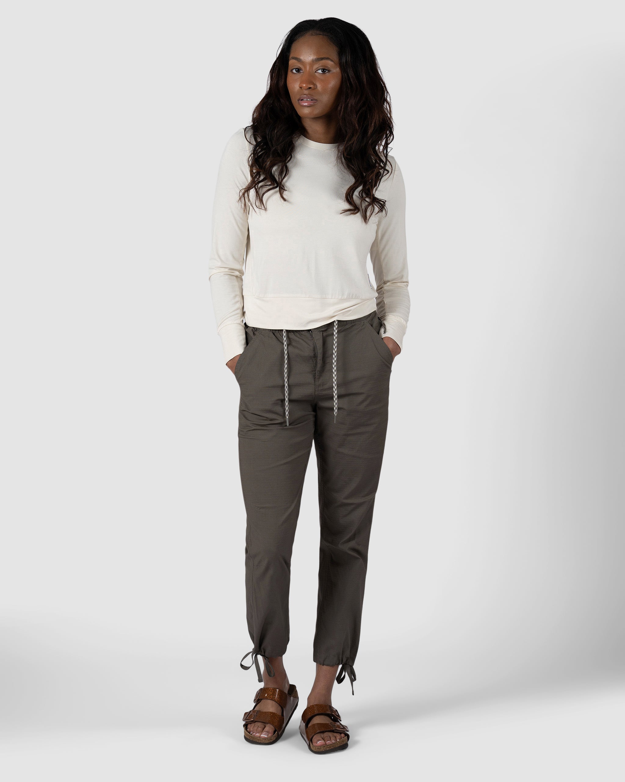 A woman with long, wavy hair stands against a plain background, hands in pockets, wearing a cream long-sleeve shirt and BEARDEDGOAT Womens Canopy Pant in olive green—crafted from durable stretch cotton with adjustable ankle drawcords.