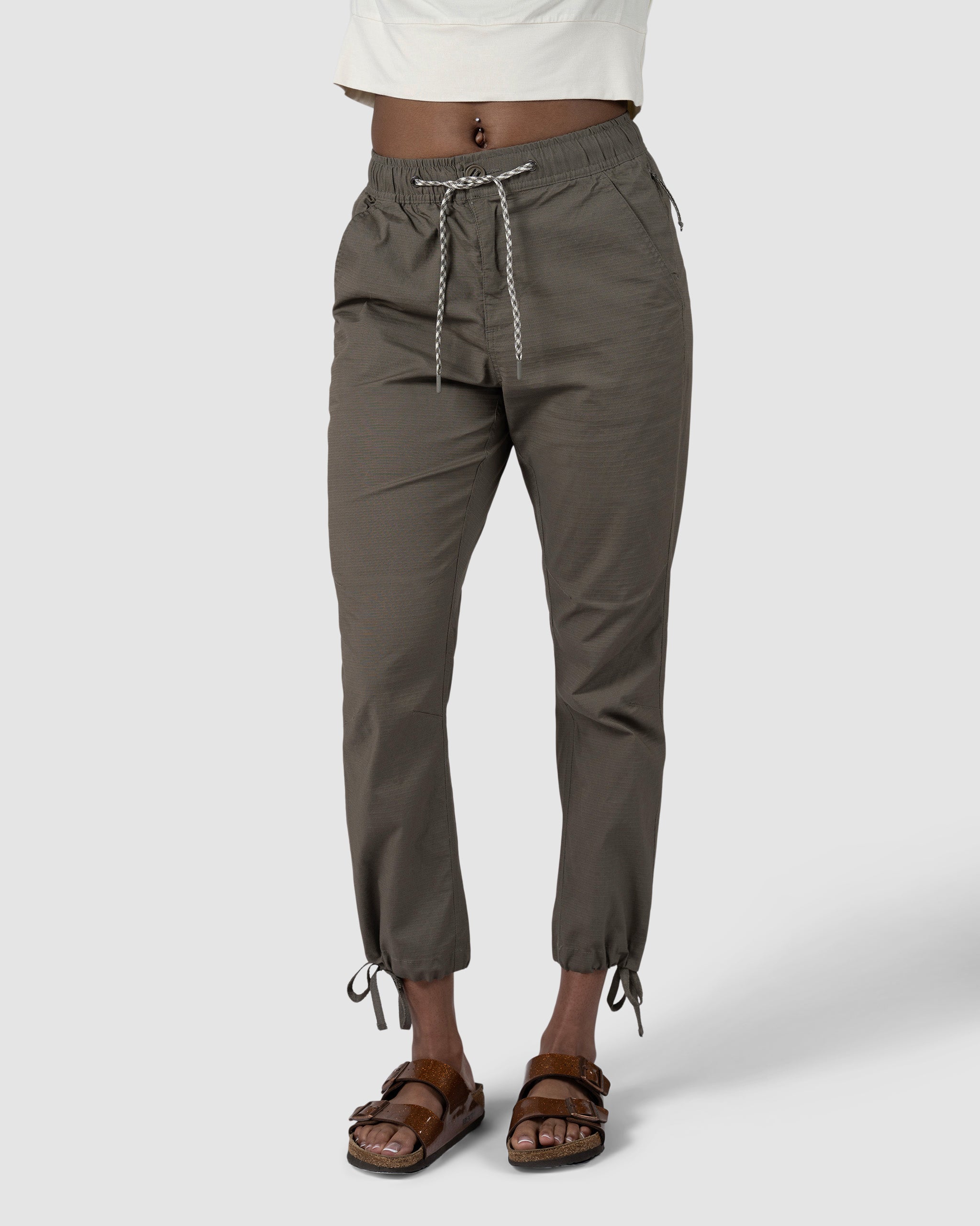 A woman wears BEARDEDGOATs Womens Canopy Pant in olive green, featuring adjustable ankle drawcords and durable stretch cotton, paired with a white cropped top and brown sandals, posing against a plain light gray background.
