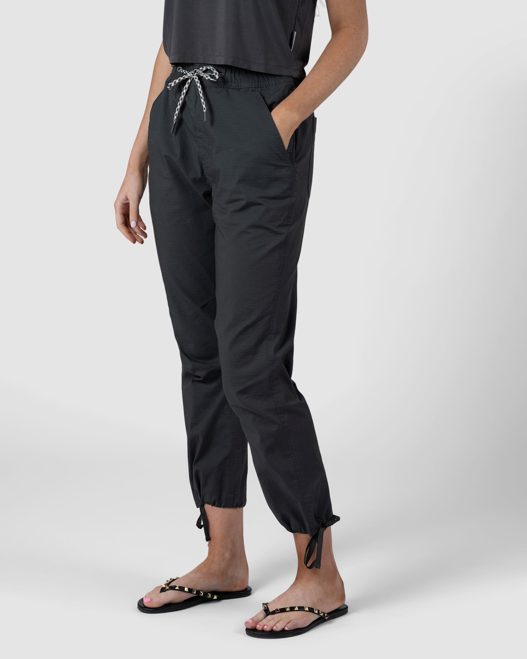 A person stands against a plain light background wearing BEARDEDGOAT Womens Canopy Pant in black, featuring adjustable ankle drawcords and side pockets, paired with black studded sandals and a dark top.