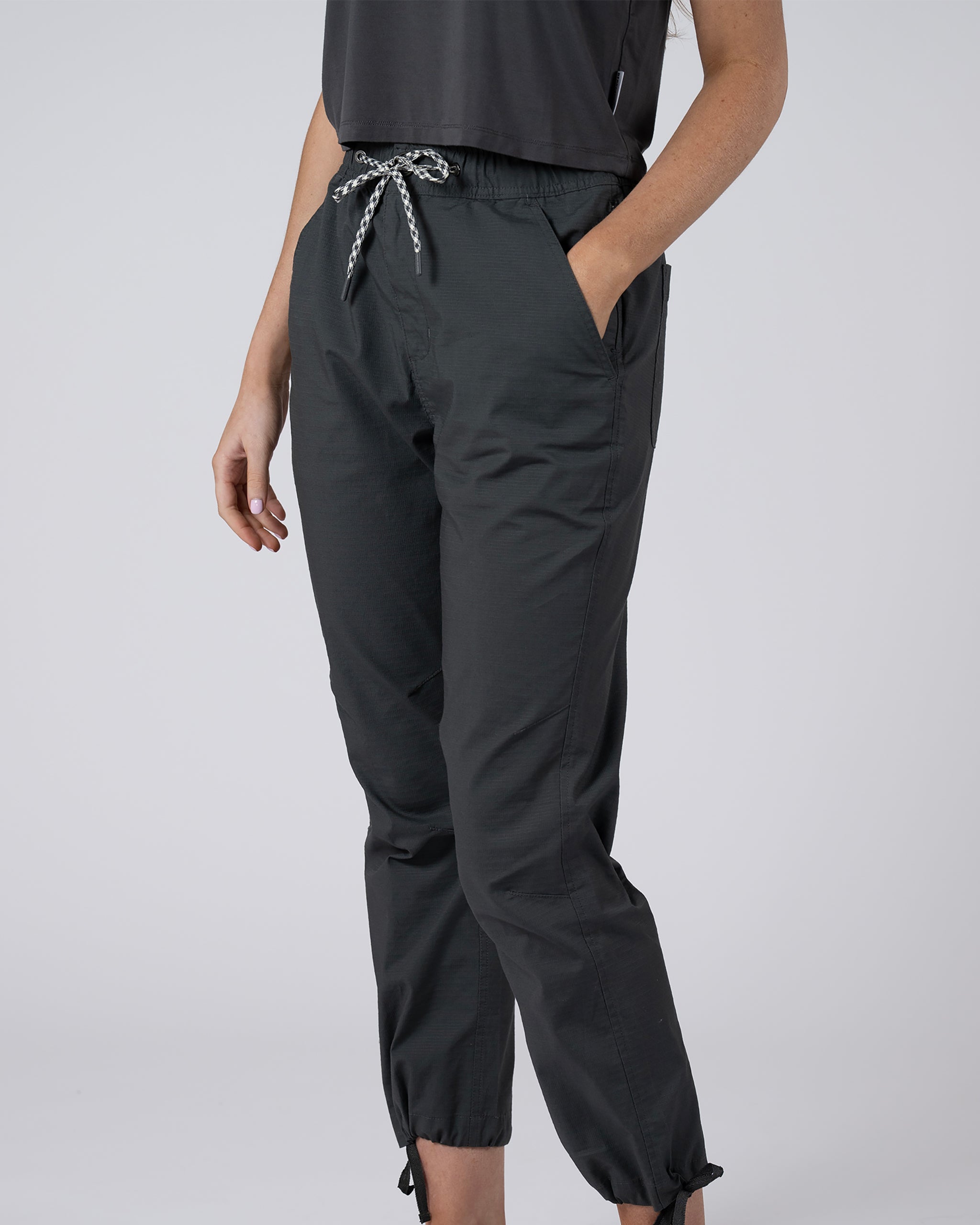 A person wears BEARDEDGOAT Women's Canopy™ Pant in dark shadow—stretch cotton ripstop with pockets and adjustable ankle drawcords—paired with a short-sleeve black top, shown from shoulders to ankles against a plain light background.
