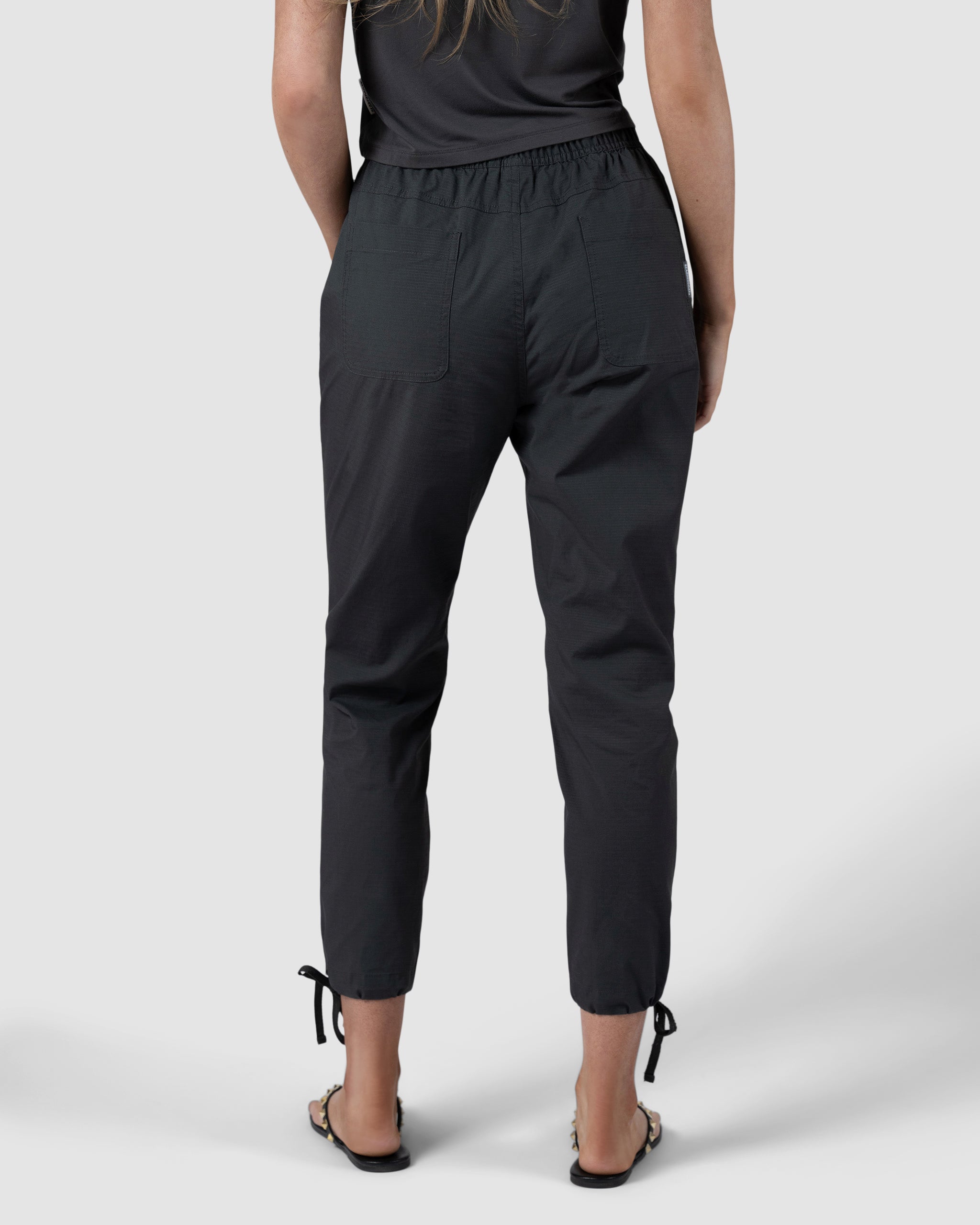 A person wearing black BEARDEDGOAT Womens Canopy Pant made from durable stretch cotton with pockets, elastic waistband, and adjustable ankle drawcords stands facing away in front of a plain light gray background.