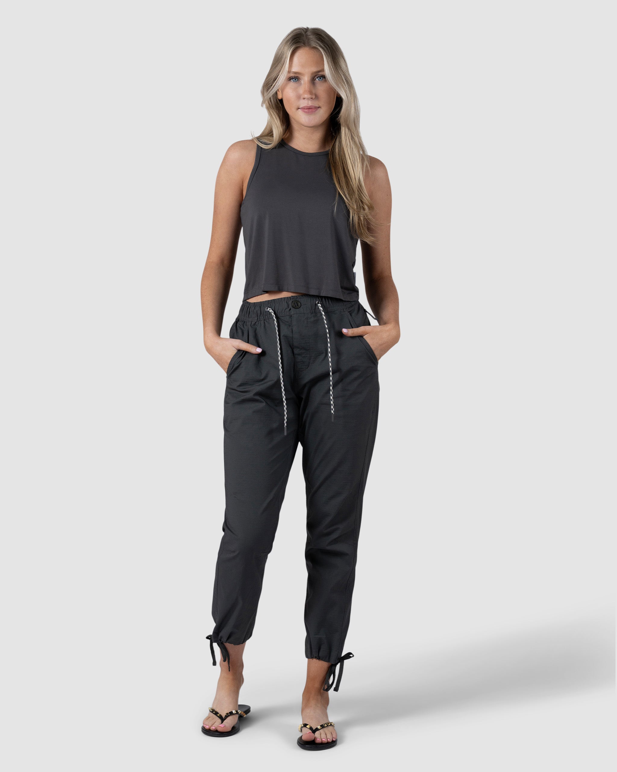 A woman with long, wavy hair poses against a plain background wearing a sleeveless black crop top, BEARDEDGOAT Womens Canopy Pant with adjustable ankle drawcords in stretch cotton, and black sandals. Her hands are in her pants pockets.