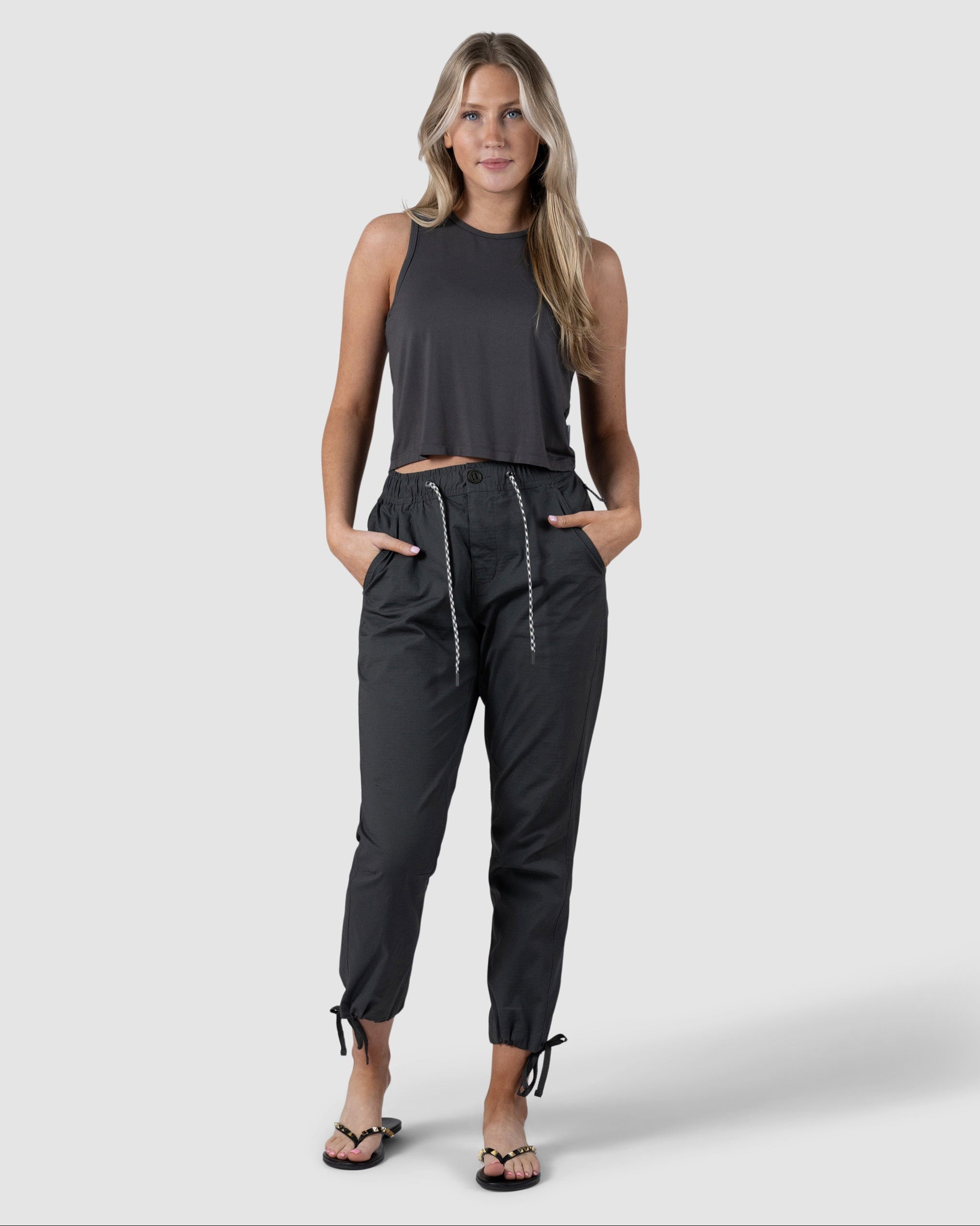 A woman with long, wavy hair poses against a plain background wearing a sleeveless black crop top, BEARDEDGOAT Womens Canopy Pant with adjustable ankle drawcords in stretch cotton, and black sandals. Her hands are in her pants pockets.