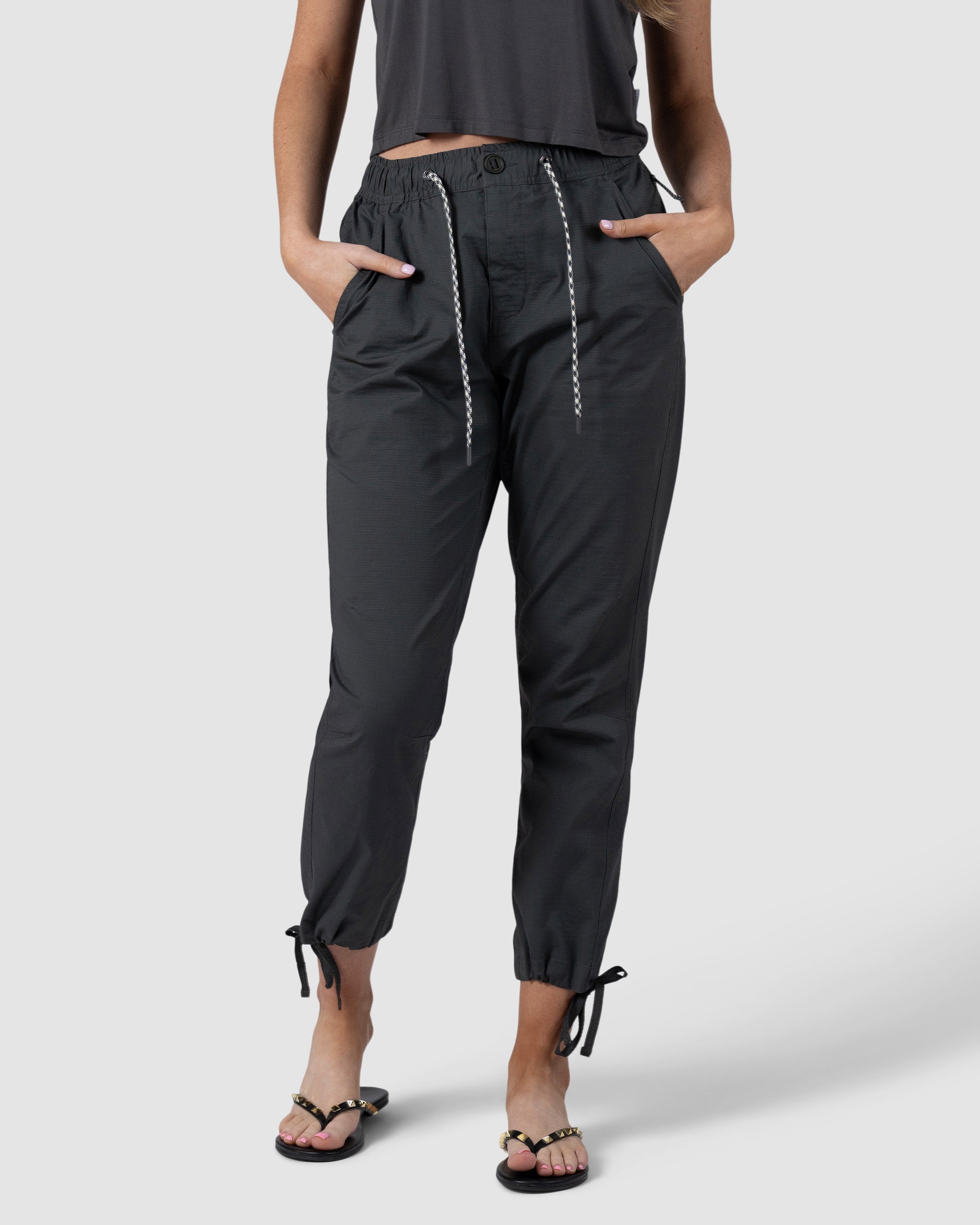 A woman models BEARDEDGOAT Womens Canopy Pant in black, crafted from durable stretch cotton with pockets and adjustable ankle drawcords, styled with a dark cropped top and black sandals featuring gold chain details against a plain background.
