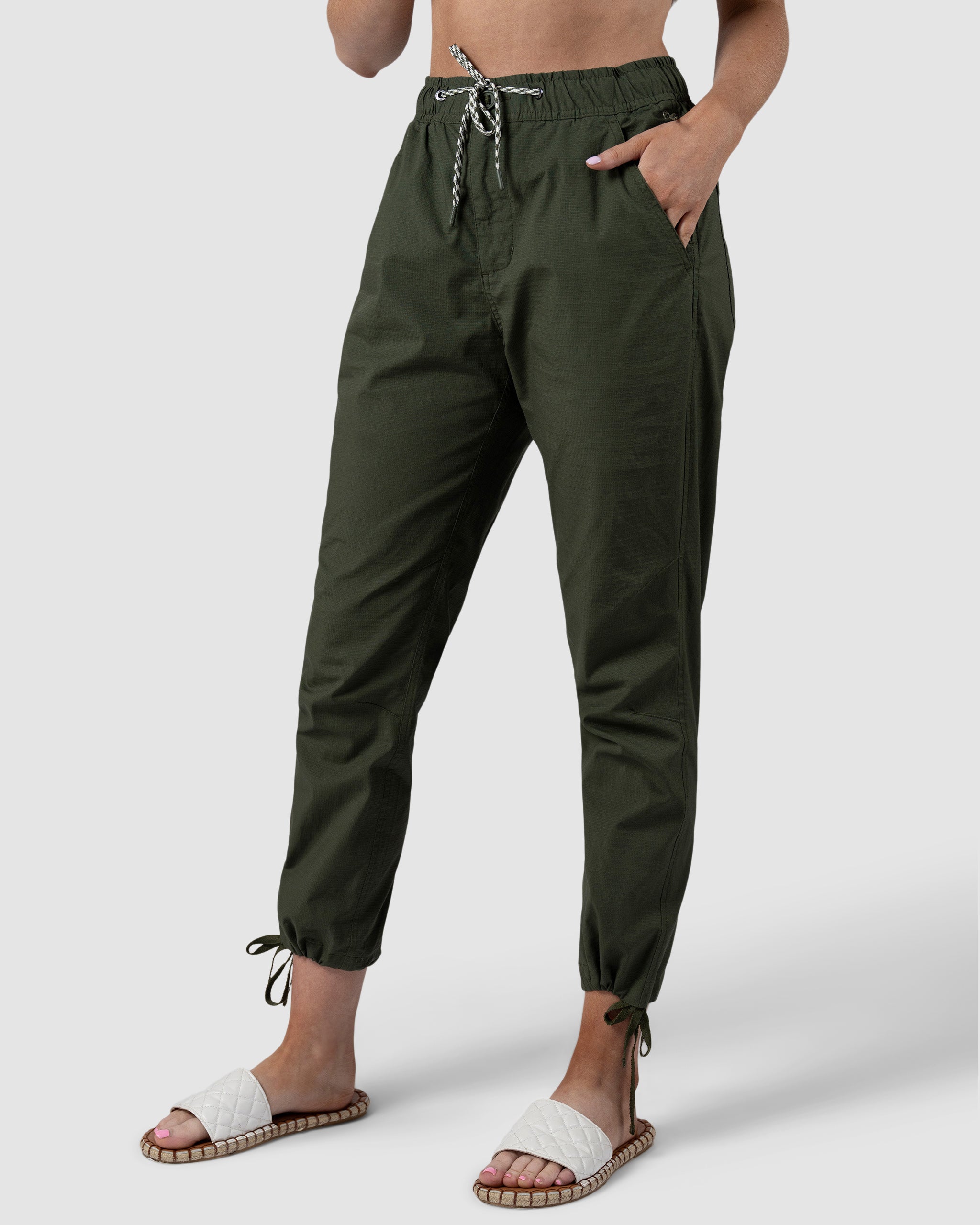A person wears BEARDEDGOAT Womens Canopy Pant in olive green, crafted from durable stretch cotton with elastic cuffs and adjustable ankle drawcords, hands in pockets, paired with white slide sandals. Only the lower half of the body is visible.