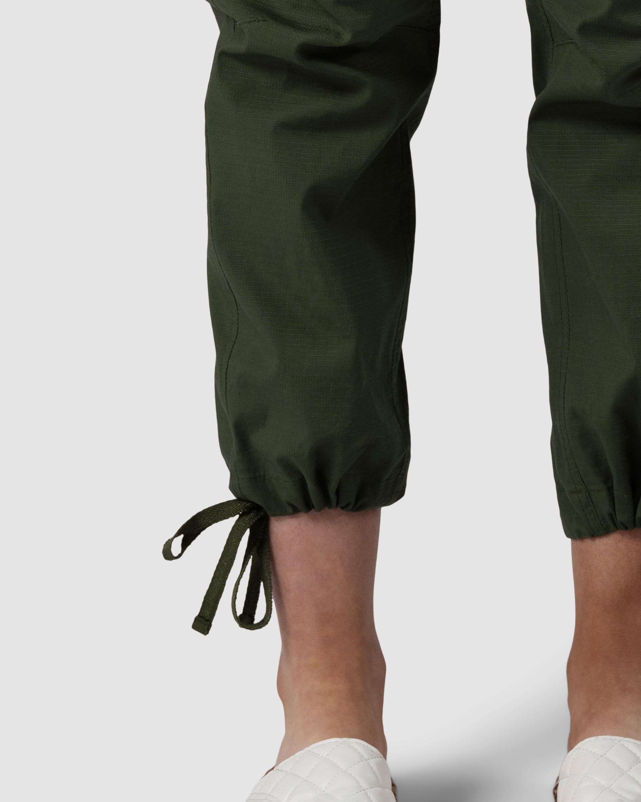 Close-up of a person wearing BEARDEDGOAT Womens Canopy Pant in durable stretch cotton with adjustable ankle drawcords, paired with white quilted slip-ons, standing against a plain light background.