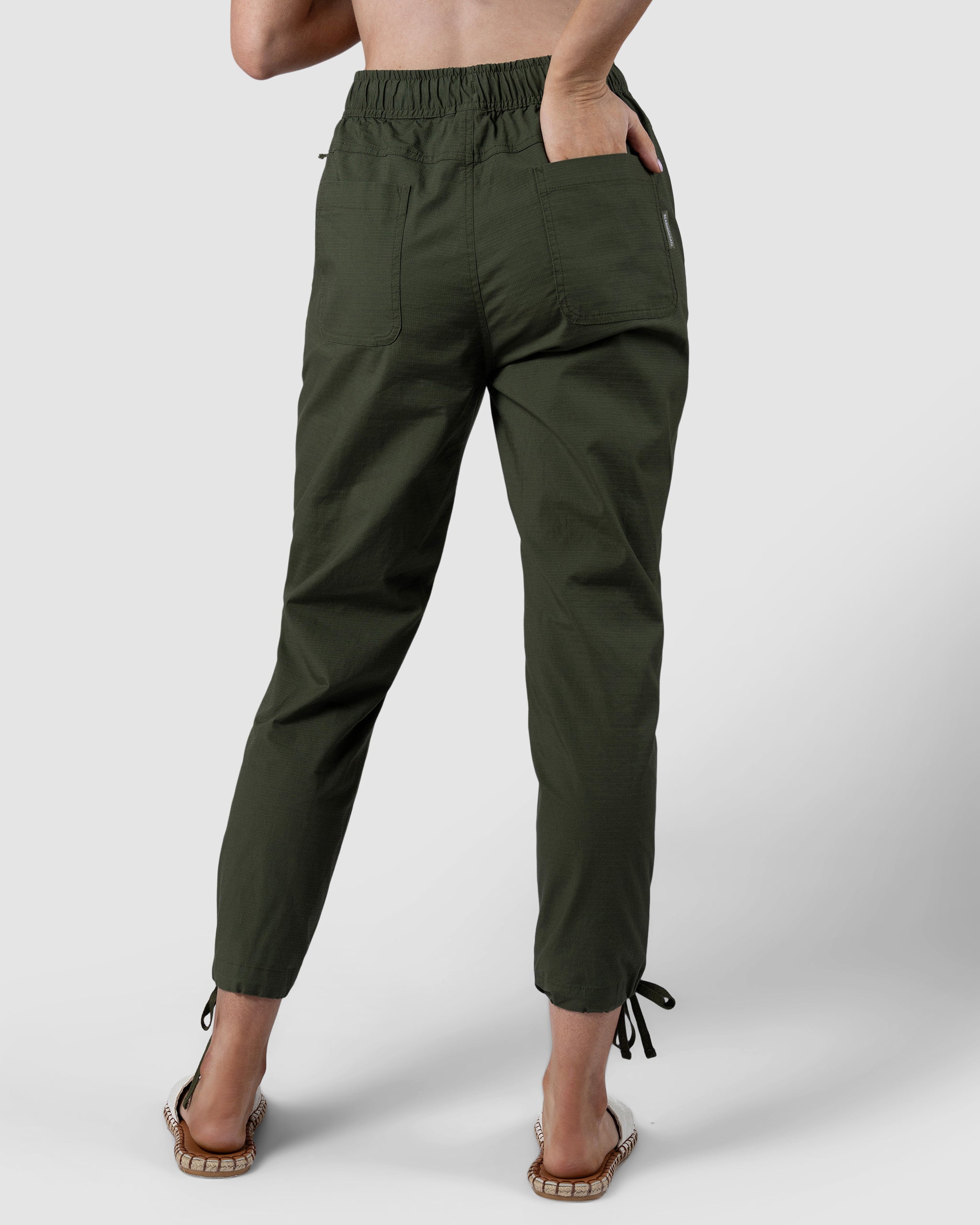 A person stands barefoot against a plain light background wearing BEARDEDGOAT Womens Canopy Pant in olive green—high-waisted, crafted from durable stretch cotton, with back pockets and adjustable ankle drawcords.