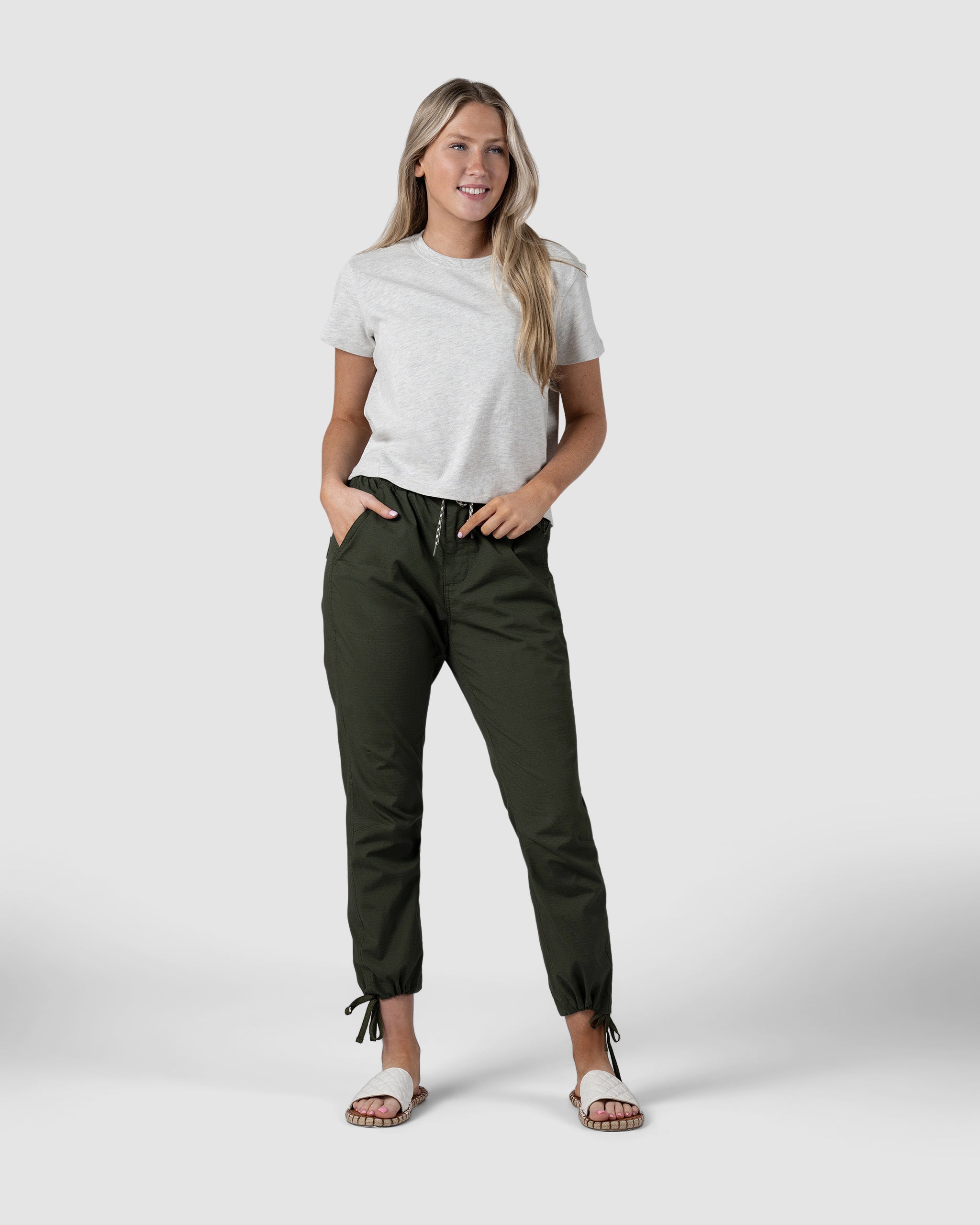 A woman stands facing forward with hands in the pockets of BEARDEDGOAT Womens Canopy Pant in olive green—crafted from durable stretch cotton with adjustable ankle drawcords—paired with a light gray t-shirt and white sandals against a plain background.
