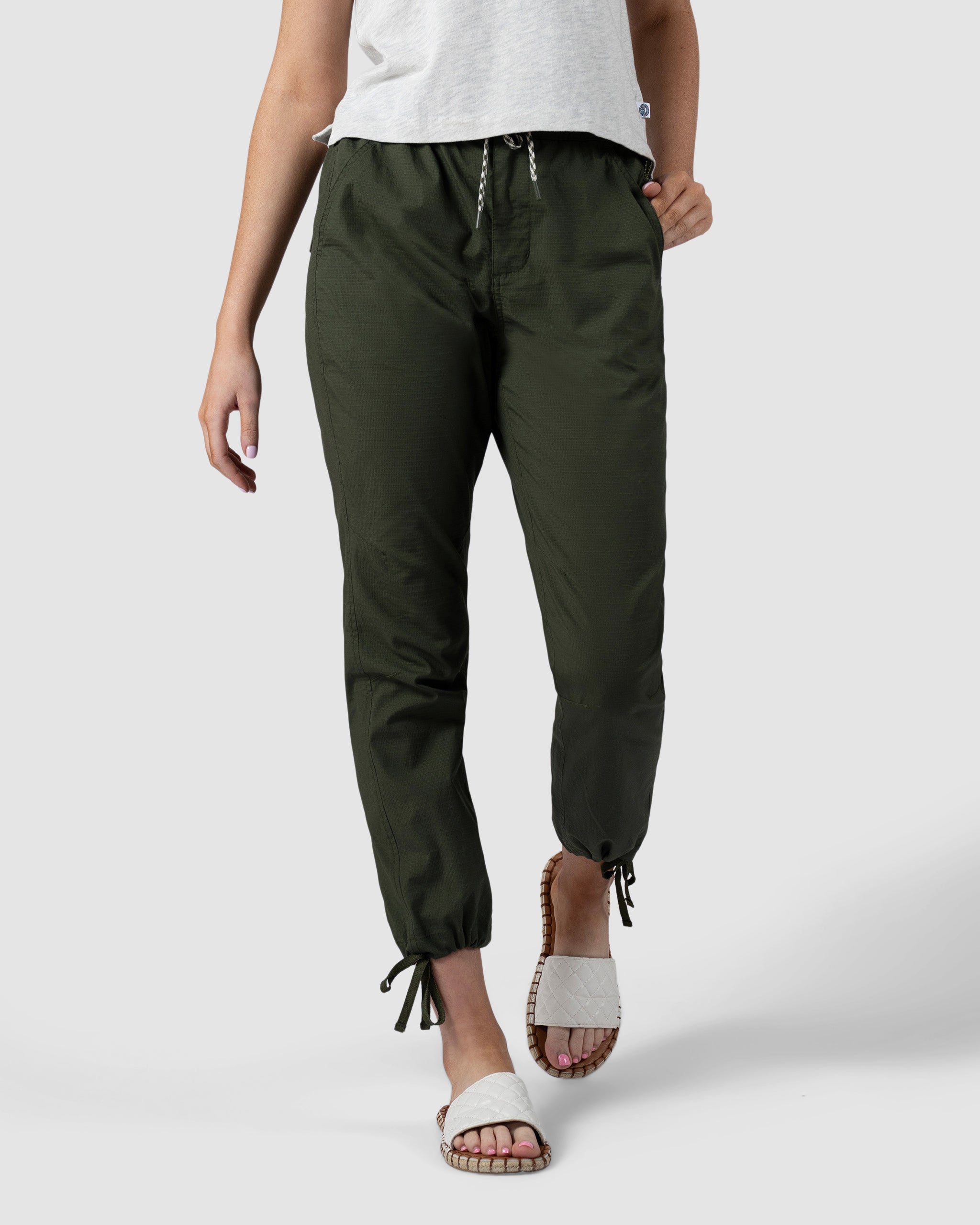 A person wears BEARDEDGOAT Womens Canopy Pant joggers in olive green, made from stretch cotton with adjustable ankle drawcords, paired with a white t-shirt and slide sandals. Only the lower half is shown against a plain light gray background.