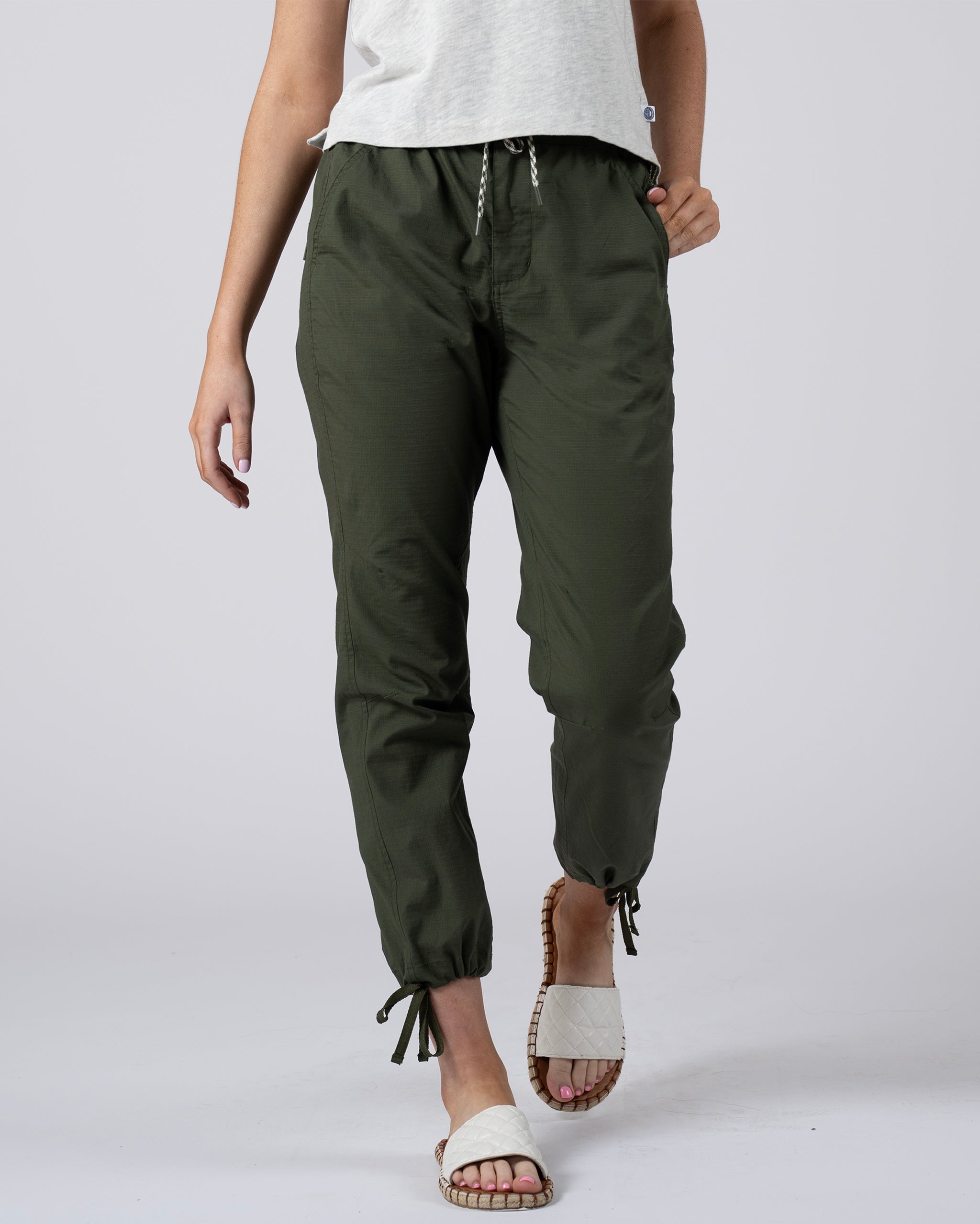 A person wearing BEARDEDGOAT Women's Canopy™ Pant in Deep Moss with adjustable ankle drawcords, a light gray top, and white slide sandals stands against a plain background. The image is cropped from the shoulders down.