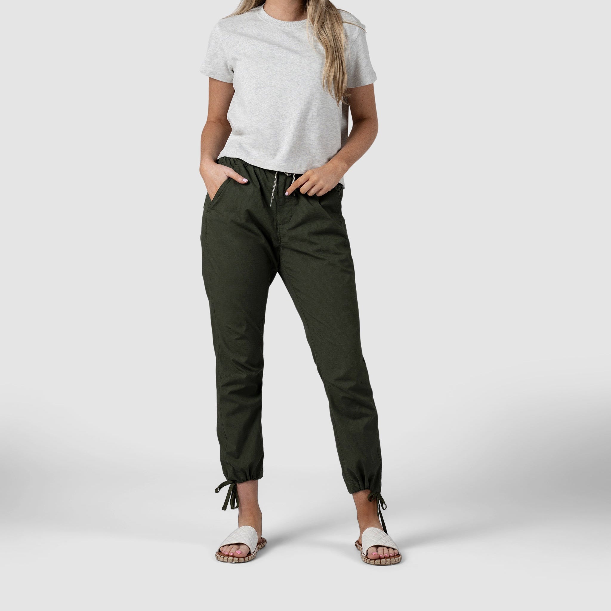 A person stands with hands in pockets against a plain light background, wearing a light gray t-shirt, BEARDEDGOAT Womens Canopy Pant in dark green with a tapered fit and ankle ties, and white sandals.