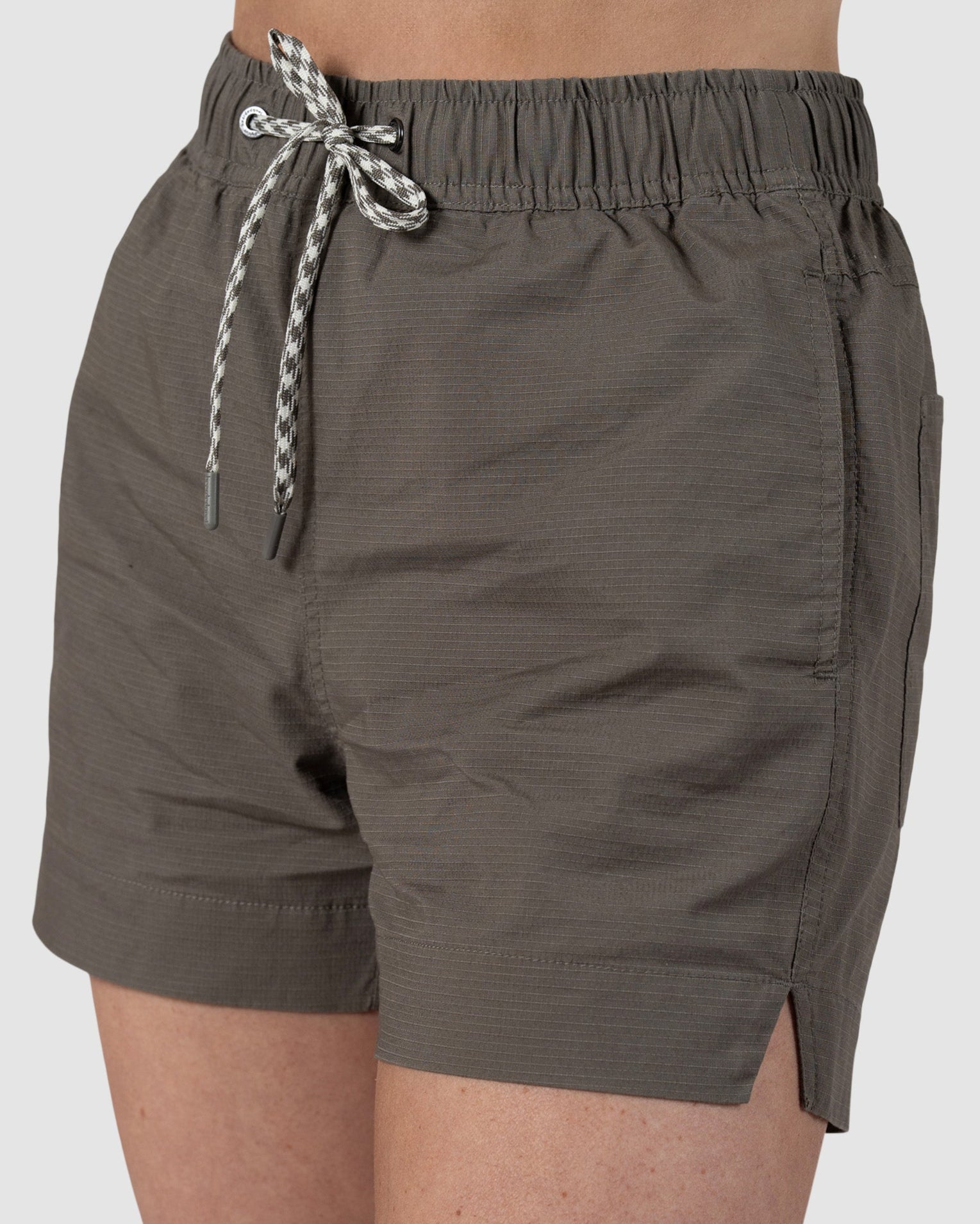 A person wears BEARDEDGOAT Womens Canopy Short in olive green, featuring an elastic waistband with a white and black drawstring, side pockets, and small side hem slits. Made from stretch cotton ripstop for breathable comfort and a relaxed fit.