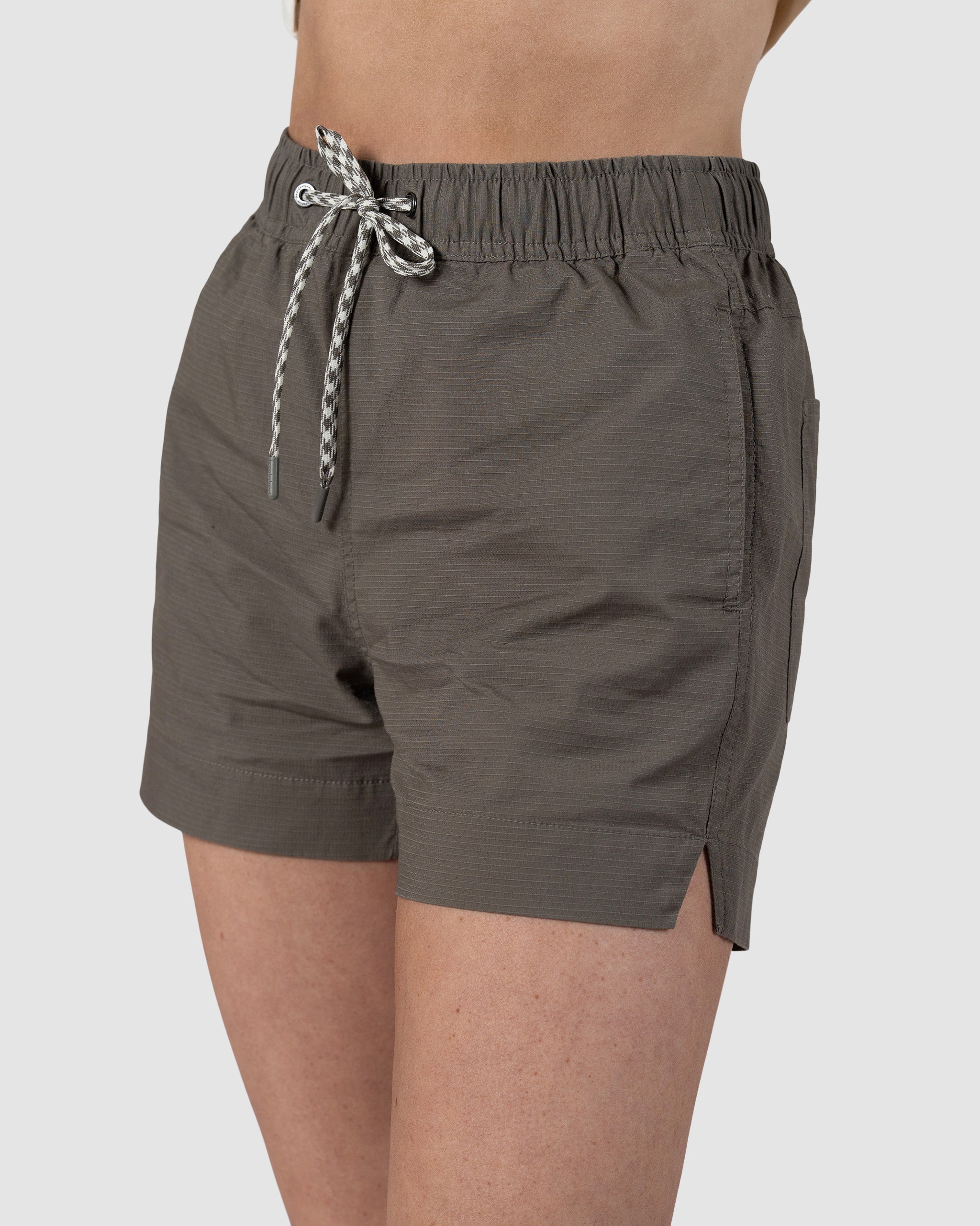 A person wears BEARDEDGOAT Womens Canopy Short in olive green, featuring an elastic waistband with a white and black drawstring, side pockets, and small side hem slits. Made from stretch cotton ripstop for breathable comfort and a relaxed fit.