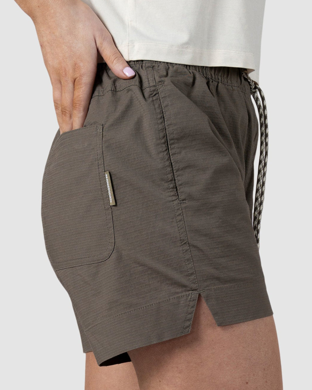 A woman wears BEARDEDGOATs Womens Canopy Short in olive green, made from stretch cotton ripstop with a side pocket and drawstring waistband, paired with a white shirt for breathable comfort.