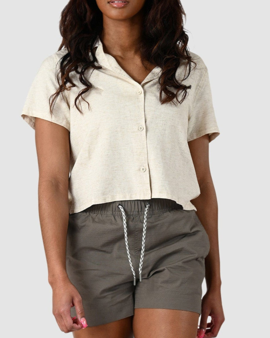 A woman stands against a plain background wearing BEARDEDGOAT’s Women’s Canopy Short in olive green stretch cotton ripstop, a short-sleeve cream button-up shirt, and brown sandals. She has long, wavy hair and a relaxed, comfortable look.