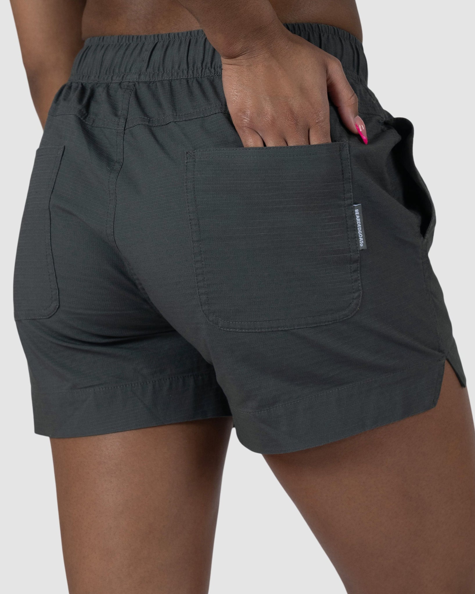 A person wears the BEARDEDGOAT Womens Canopy Short in dark green stretch cotton ripstop with an elastic waistband, standing against a light gray background. Only the lower torso and upper legs are shown; one hand is in a back pocket.