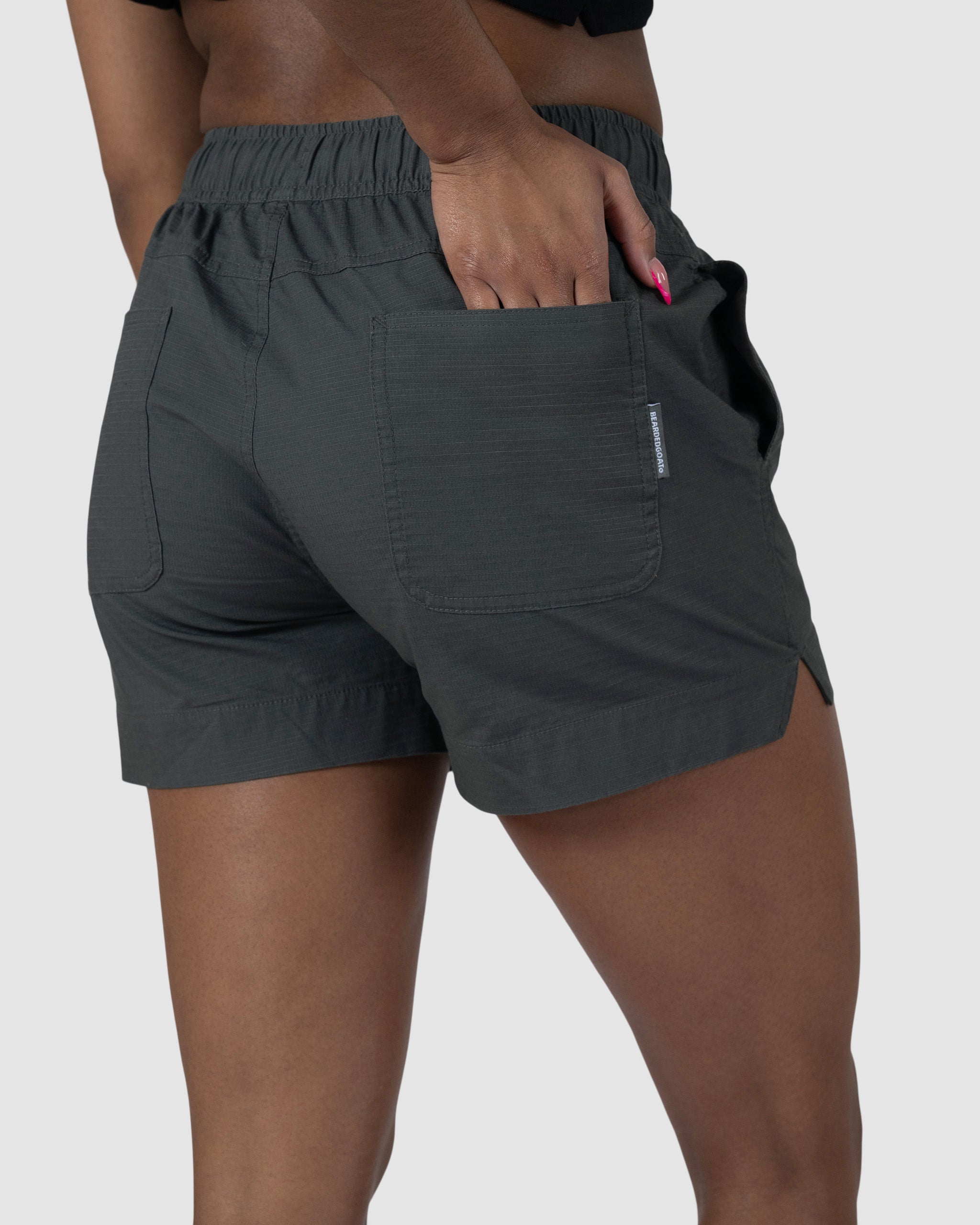 A person wears the BEARDEDGOAT Womens Canopy Short in dark green stretch cotton ripstop with an elastic waistband, standing against a light gray background. Only the lower torso and upper legs are shown; one hand is in a back pocket.