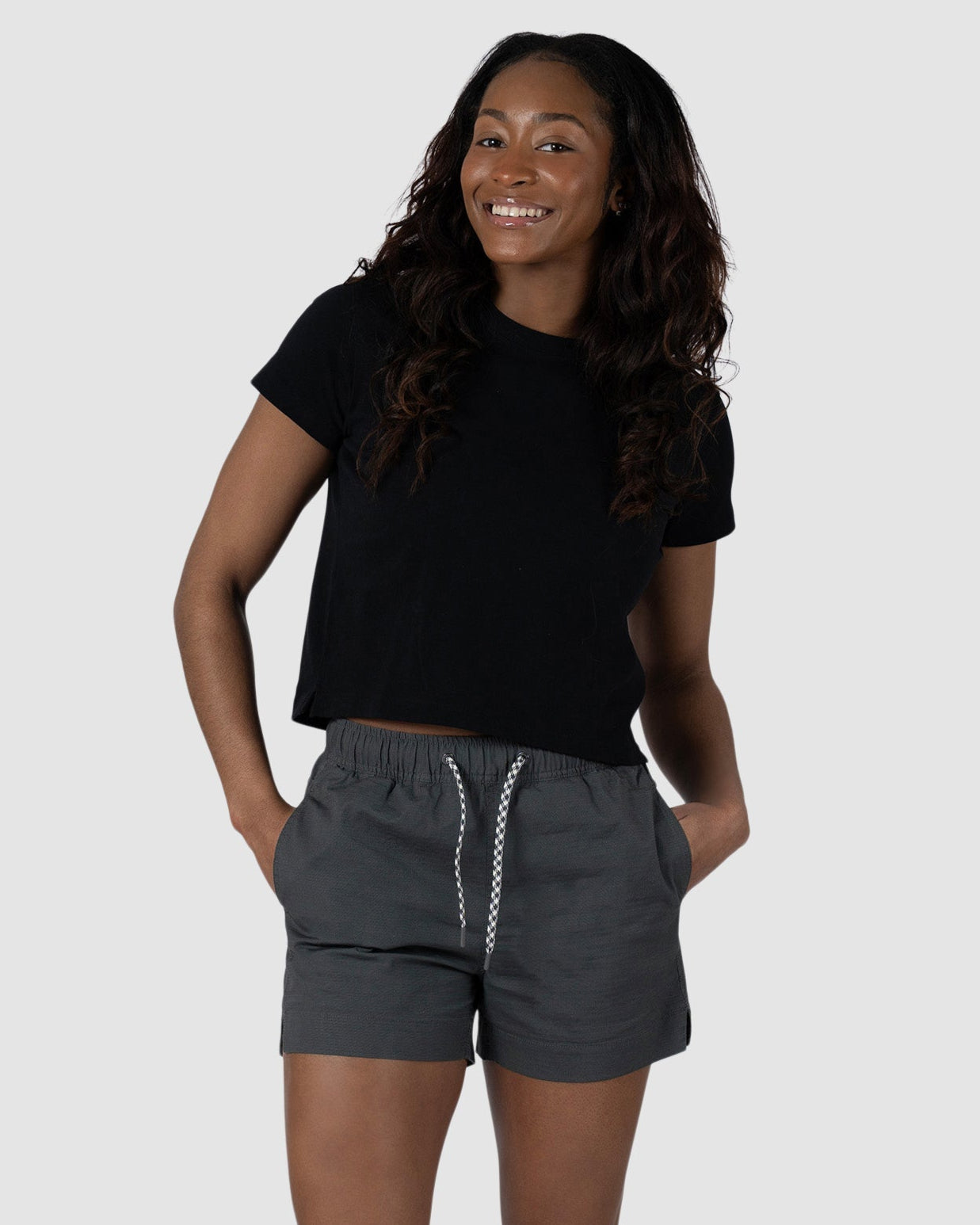 A woman smiles with her hands in the pockets of her BEARDEDGOAT Womens Canopy Short, made from stretch cotton ripstop for comfort. She pairs it with a black t-shirt and brown sandals, standing against a plain light gray background.