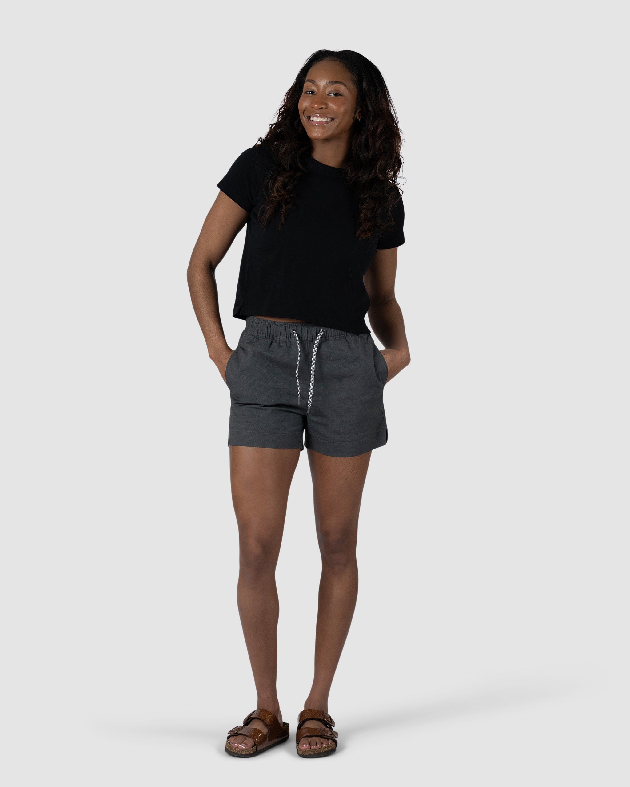 A woman smiles with her hands in the pockets of her BEARDEDGOAT Womens Canopy Short, made from stretch cotton ripstop for comfort. She pairs it with a black t-shirt and brown sandals, standing against a plain light gray background.