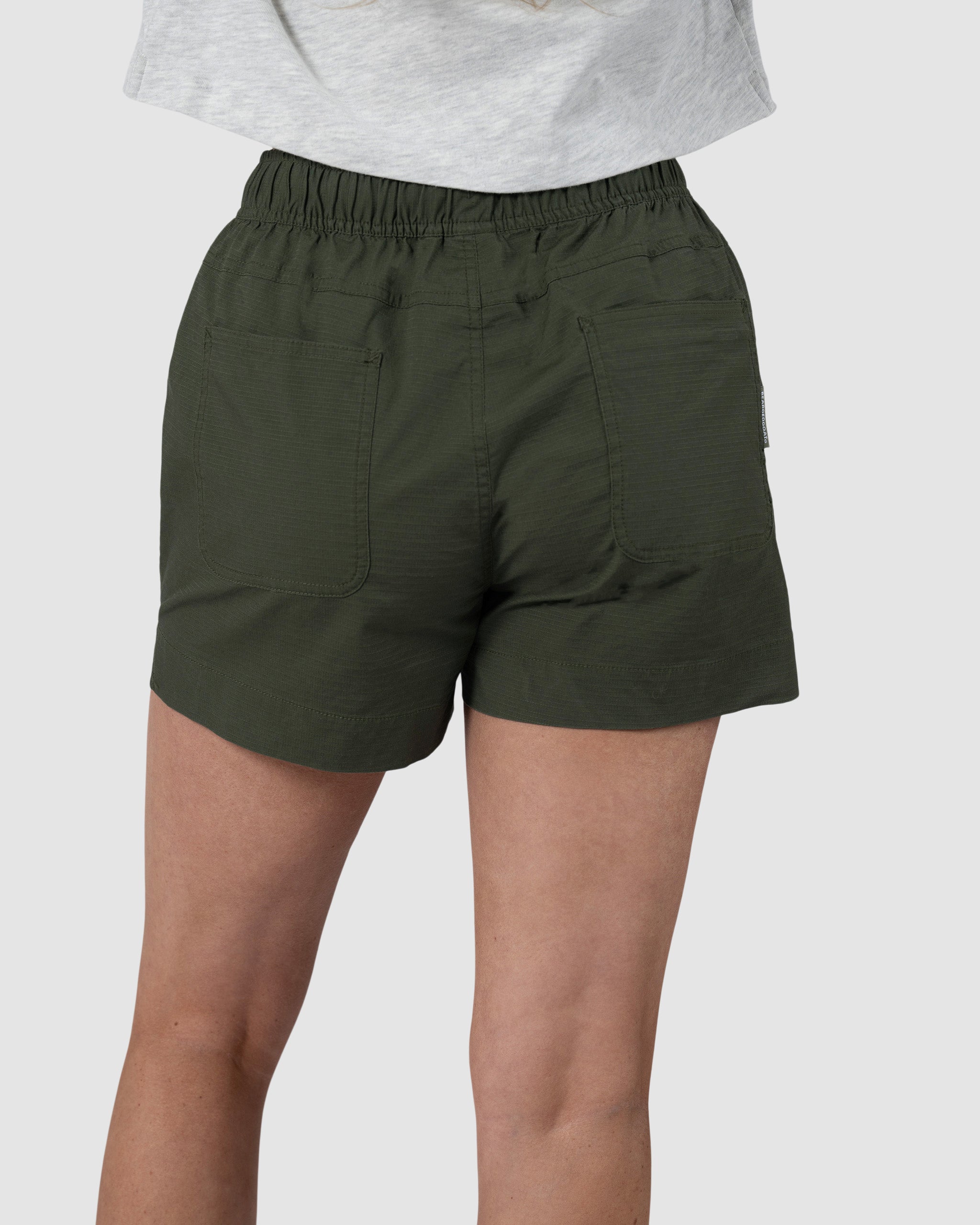 A person wearing BEARDEDGOAT Womens Canopy Short in olive green—crafted from stretch cotton ripstop with an elastic waistband and two large back pockets—is shown waist-down from behind, paired with a light gray shirt.