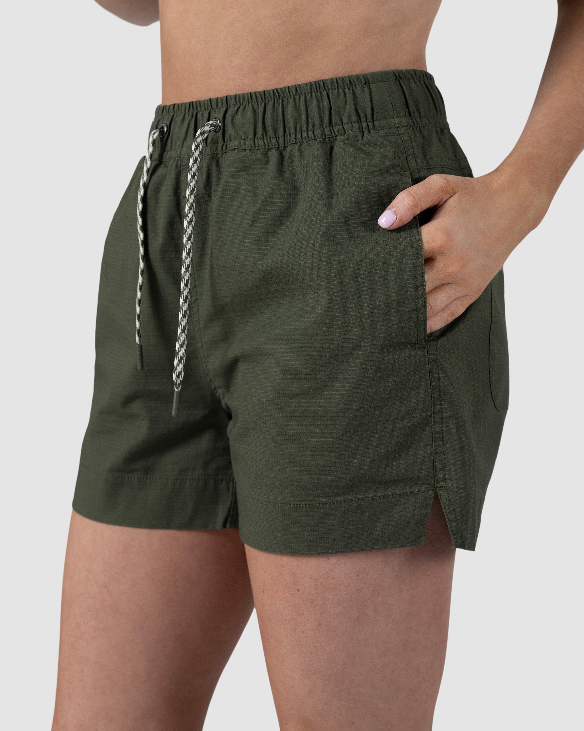 A person wears Womens Canopy Short by BEARDEDGOAT in olive green stretch cotton ripstop, with an elastic waistband and side pockets. Only their lower torso and upper thighs are visible against a plain light background.