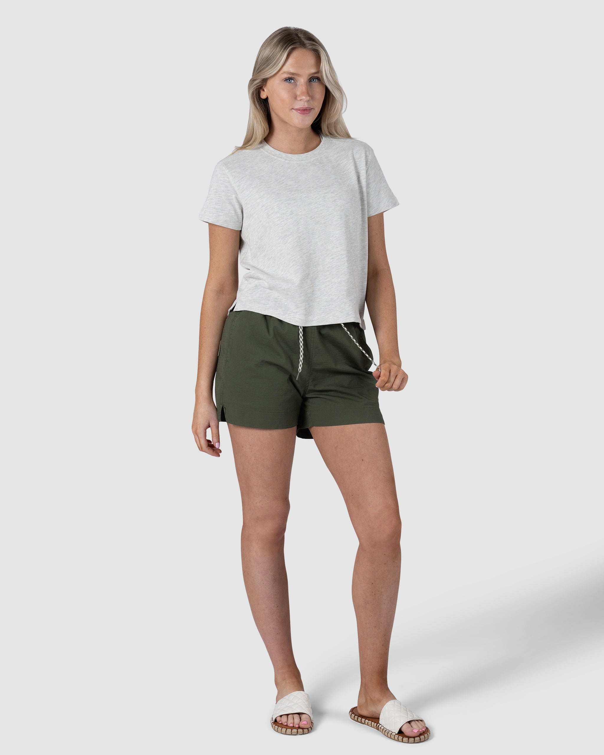 A woman with light hair and a relaxed expression stands against a plain white background, wearing a light gray tee and BEARDEDGOAT Womens Canopy Short in dark green stretch cotton ripstop, styled with white slide sandals.
