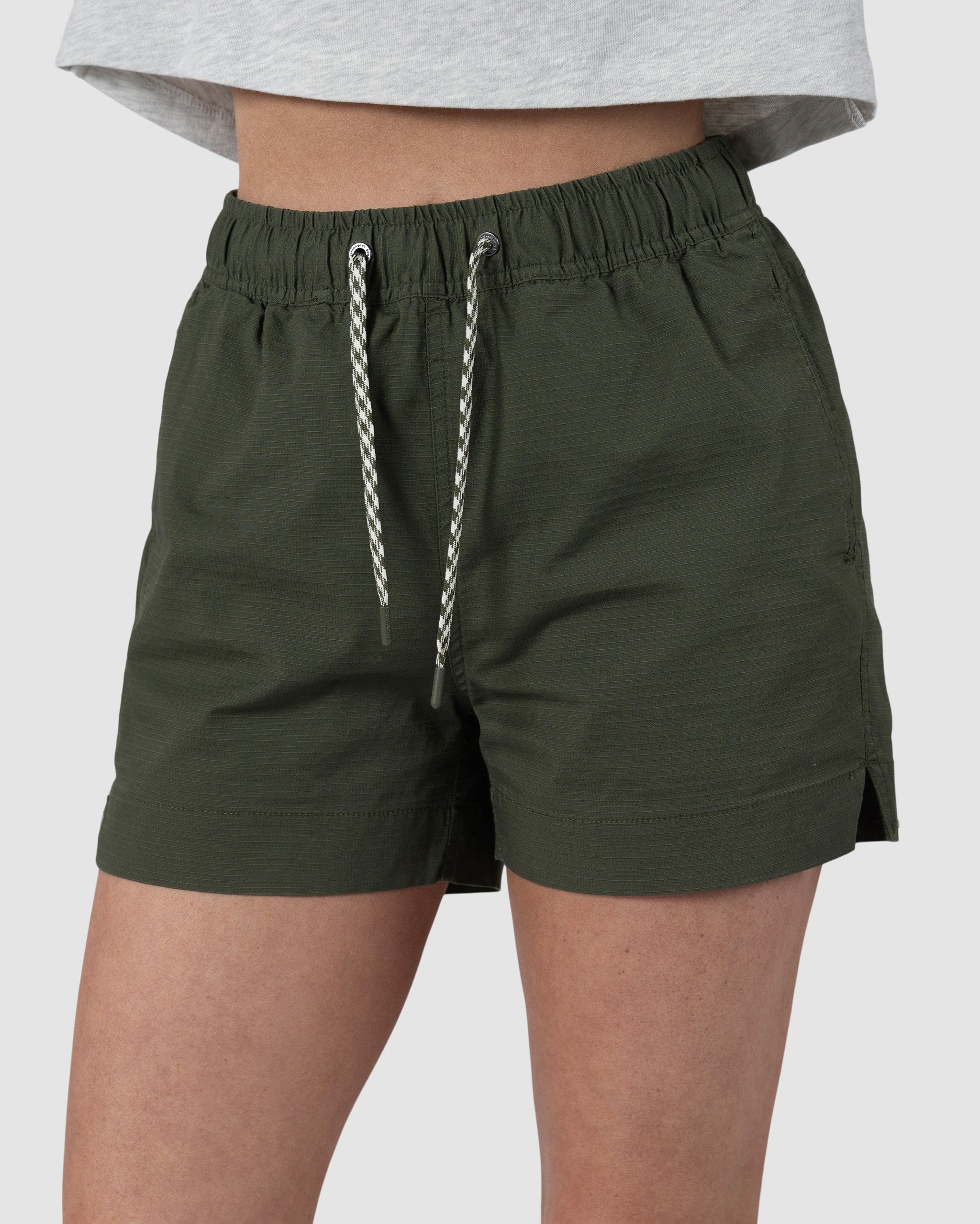 A woman wears BEARDEDGOATs Womens Canopy Short in olive green stretch cotton ripstop with a light gray shirt, shown waist to mid-thigh against a plain background, highlighting the short’s breathable comfort.