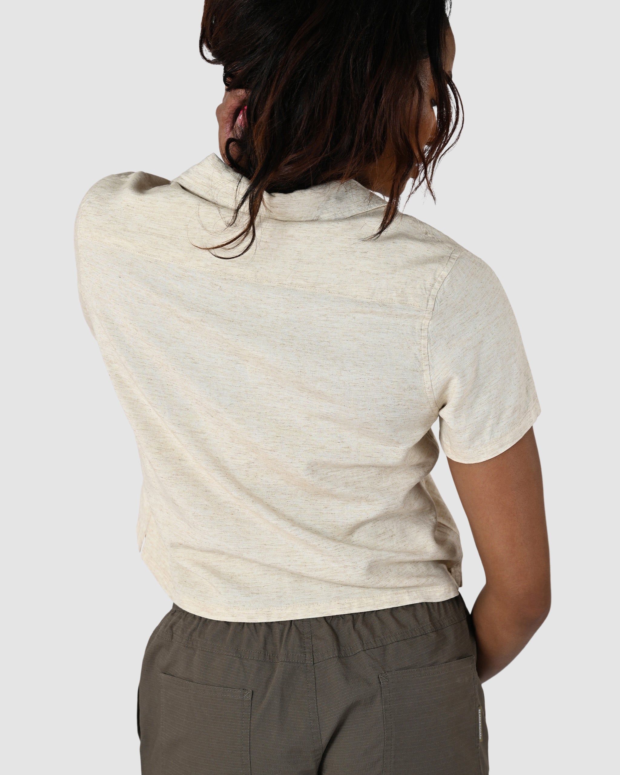 A person with long, dark hair faces away, wearing the BEARDEDGOAT Womens Breeze Shirt—short-sleeved and lightweight linen—with olive green pants, one hand at their neck, set against a plain light background.