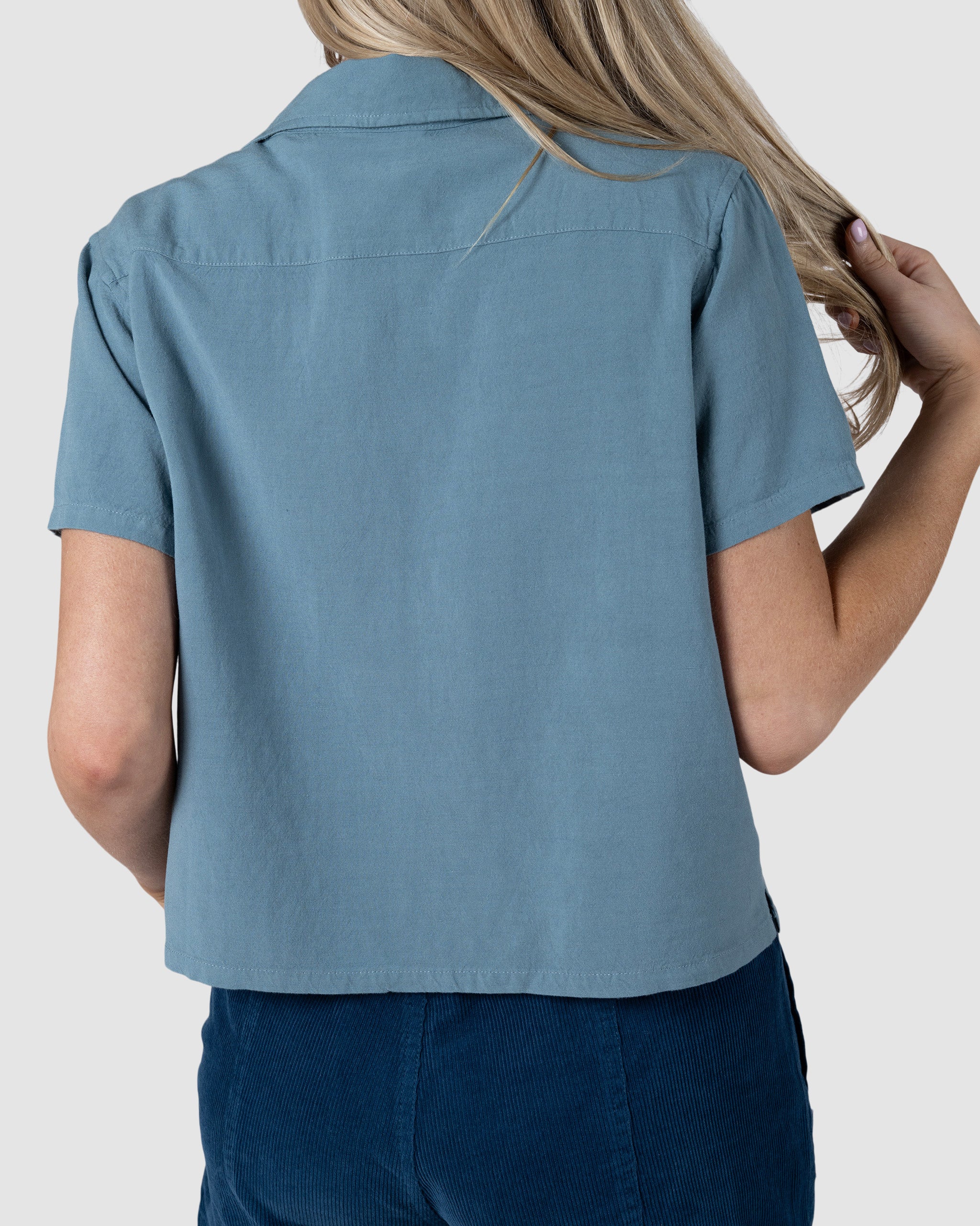 A woman with long blonde hair, shown from the back against a plain gray background, wears BEARDEDGOAT’s Women’s Breeze Shirt in light blue linen and dark blue pants, holding a section of her hair with her right hand.