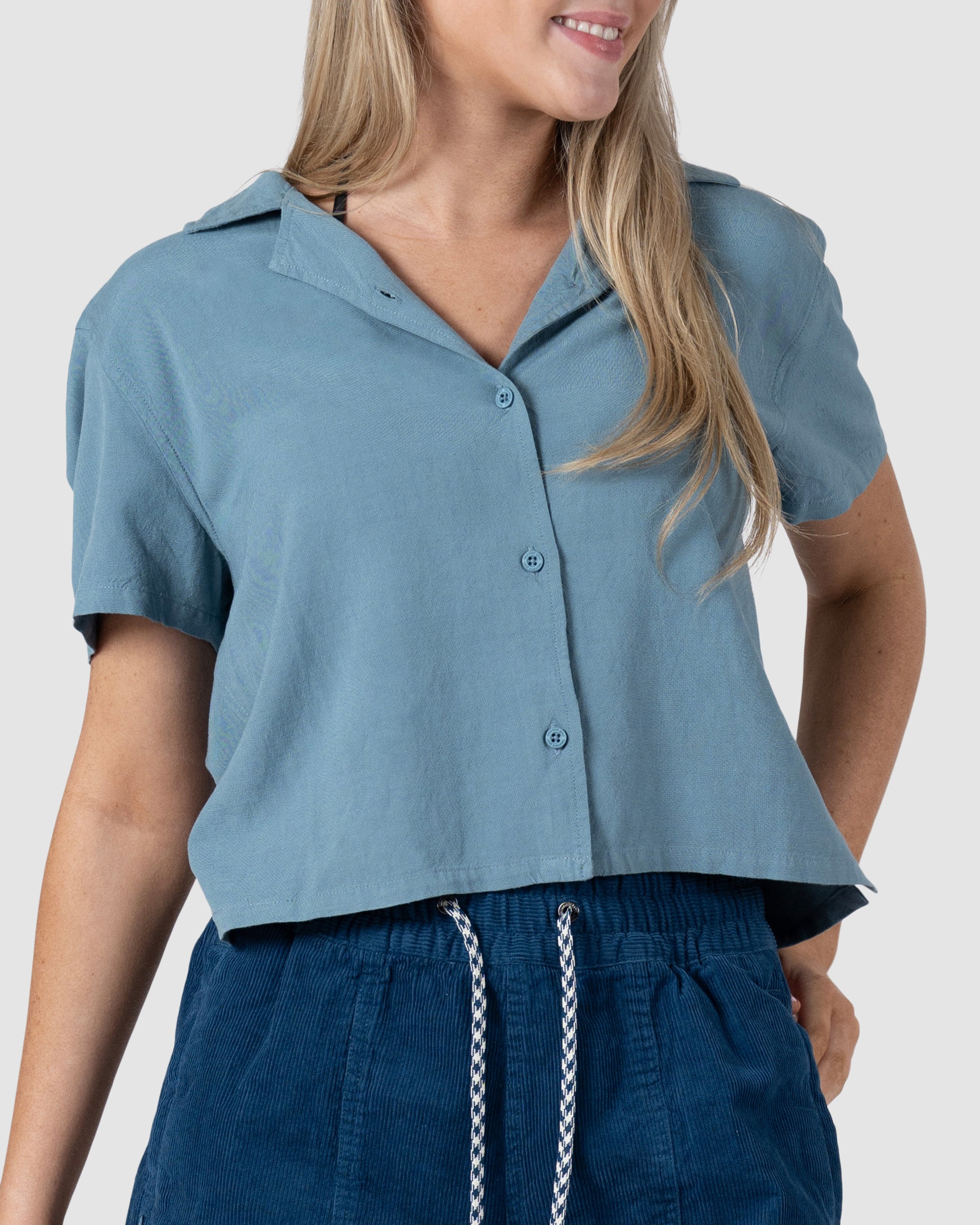 A woman models the BEARDEDGOAT Womens Breeze Shirt, a lightweight linen top with a button-front design, paired with dark blue drawstring shorts, smiling against a plain light background.