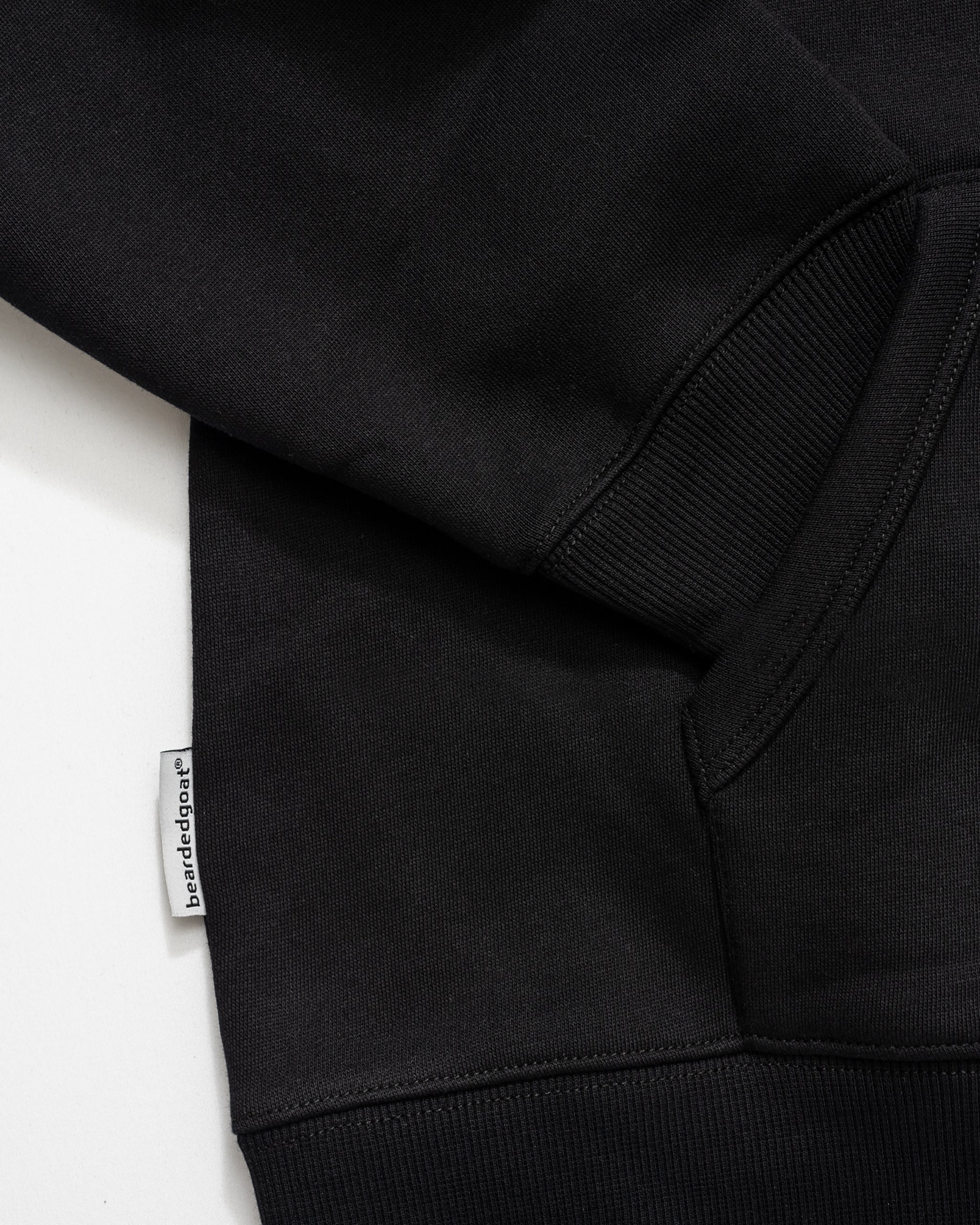 Close-up of the Men's Void Hoodie in black by beardedgoat, featuring ribbed cuffs, a front pocket, and a small white beardedgoat tag sewn into the seam near the lower edge, emphasizing the hoodie's durability.