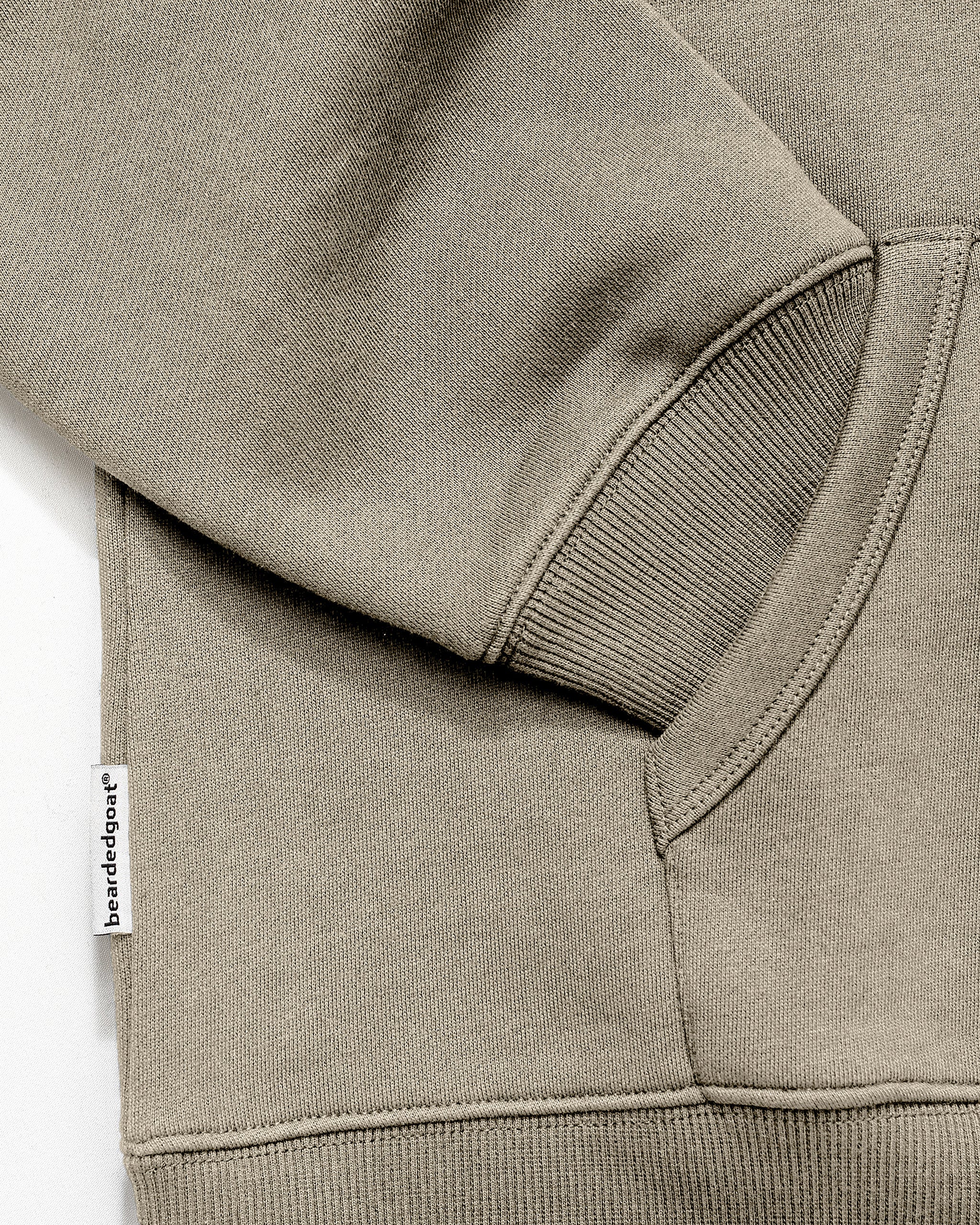 Close-up of the Men's Void Hoodie by beardedgoat in khaki, featuring heavyweight cotton, ribbed cuff, pocket, sleeve detail, and a small white "banded together" tag sewn into the side seam.