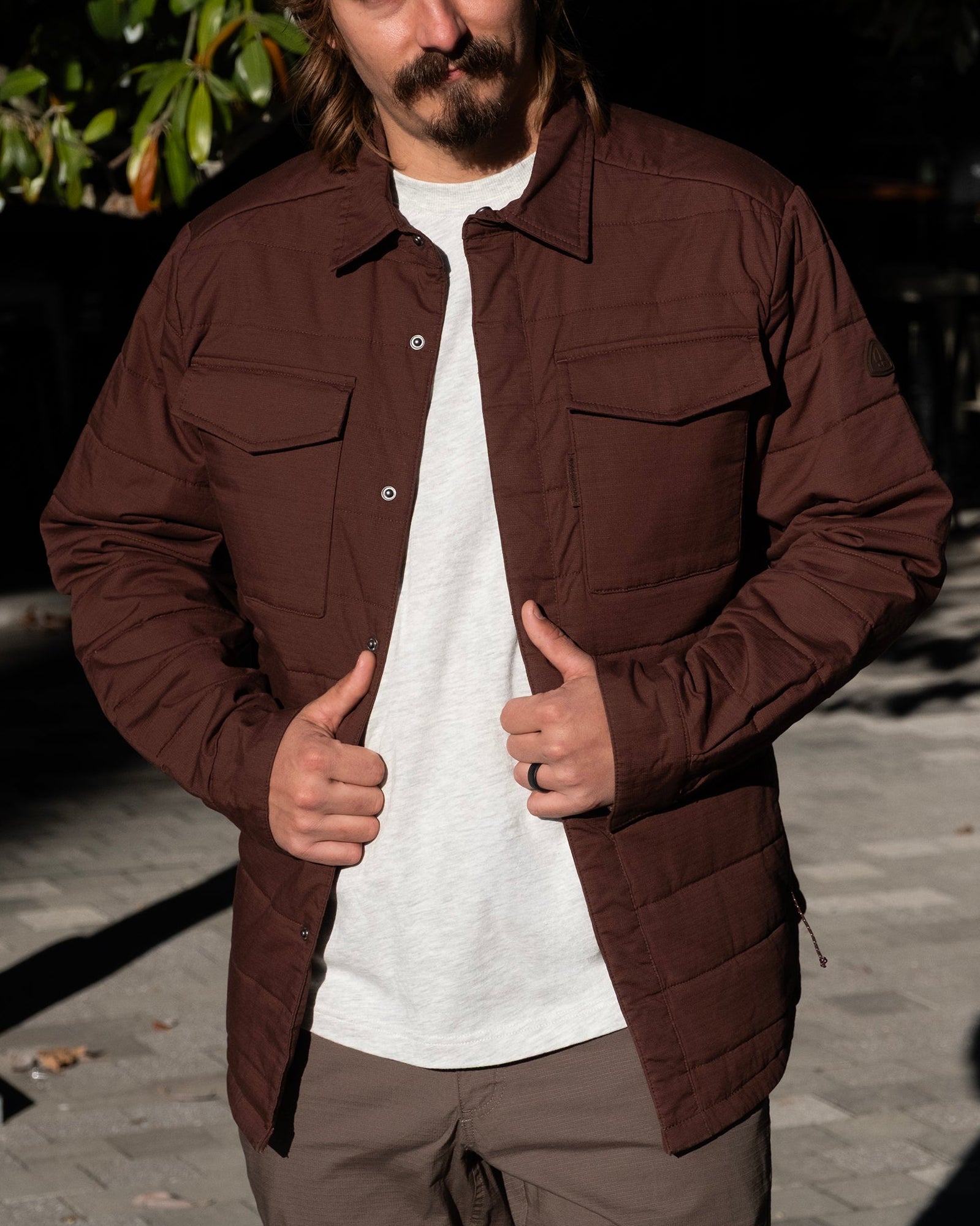 A man with long hair and a mustache wears the BEARDEDGOAT NEW — CANOPY™ Shacket in Chickory over a white shirt, standing outdoors on a sunlit paved area with trees in the background.
