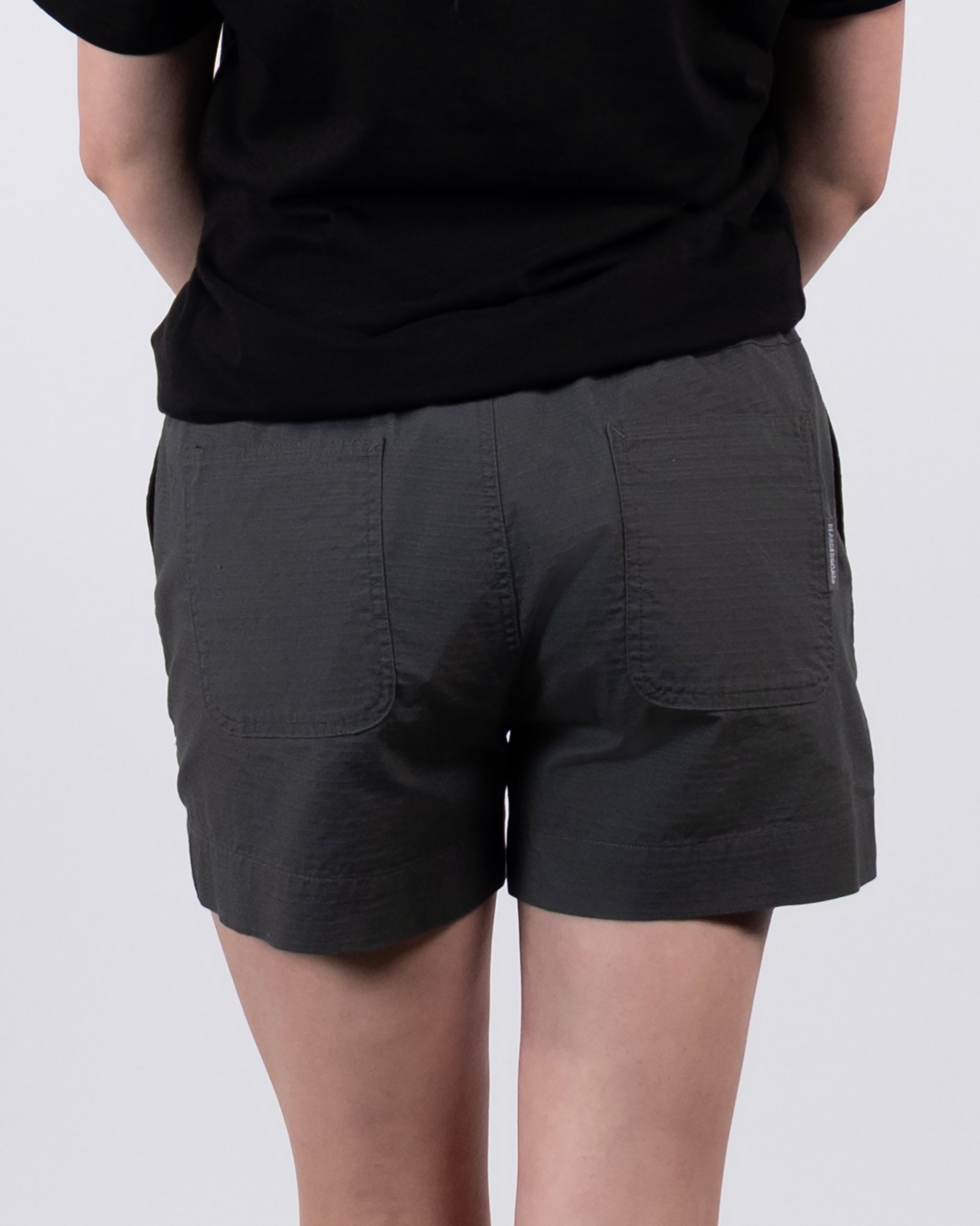 A person wearing dark gray BEARDEDGOAT Women's Canopy Shorts and a black stretch cotton ripstop T-shirt stands with their hands behind their back, facing away from the camera.