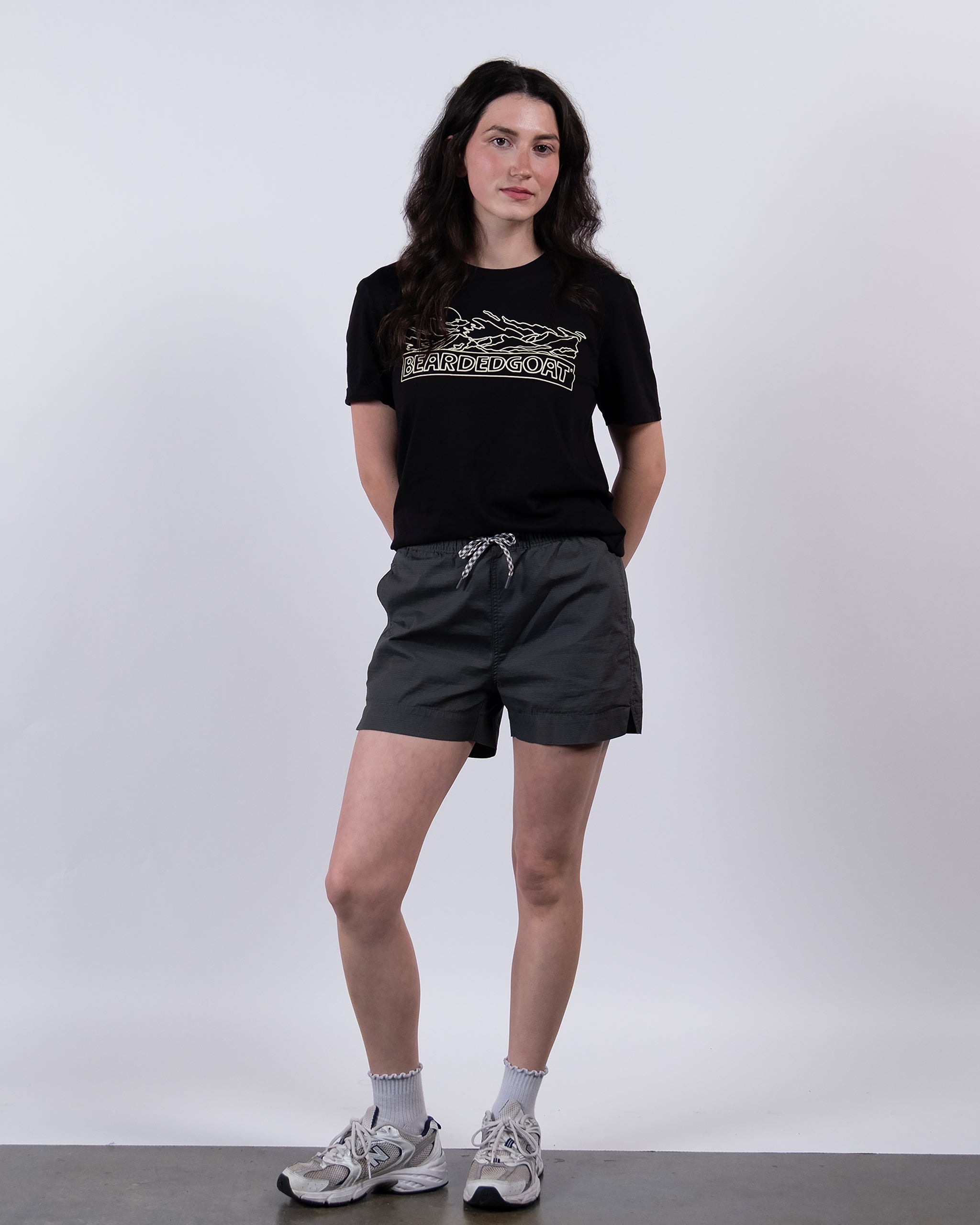A woman stands against a white background wearing BEARDEDGOAT Women's Canopy Short in dark gray stretch cotton ripstop with a patterned drawstring, paired with a black graphic tee, white socks, and gray sneakers. She looks at the camera with her hands behind her back.