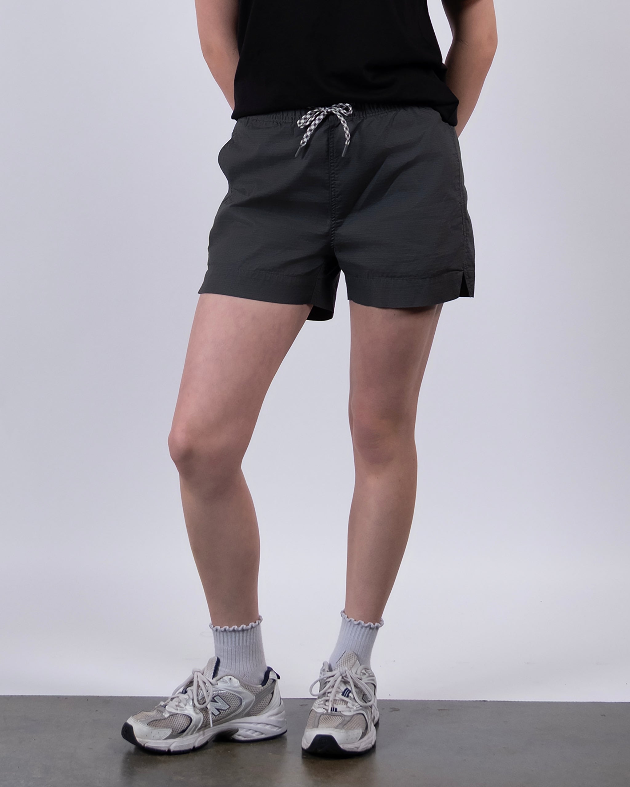 A person stands against a plain background wearing the BEARDEDGOAT Women's Canopy Short in dark gray stretch cotton ripstop, a black t-shirt, ruffled-edge white socks, and gray sneakers. Their arms are behind their back; face is not shown.