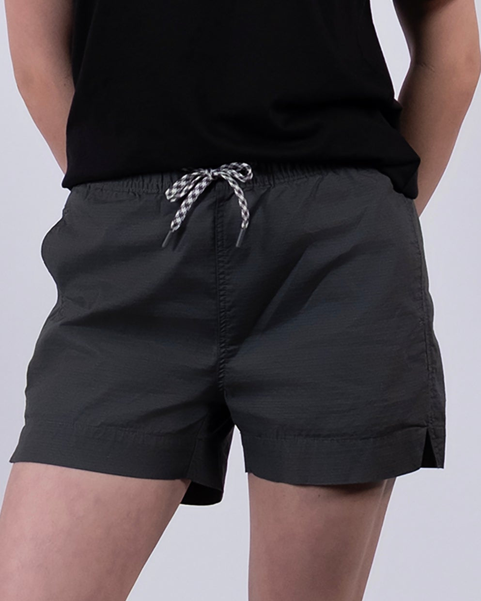 A person stands against a plain background wearing the BEARDEDGOAT Women's Canopy Short in dark gray stretch cotton ripstop, a black t-shirt, ruffled-edge white socks, and gray sneakers. Their arms are behind their back; face is not shown.