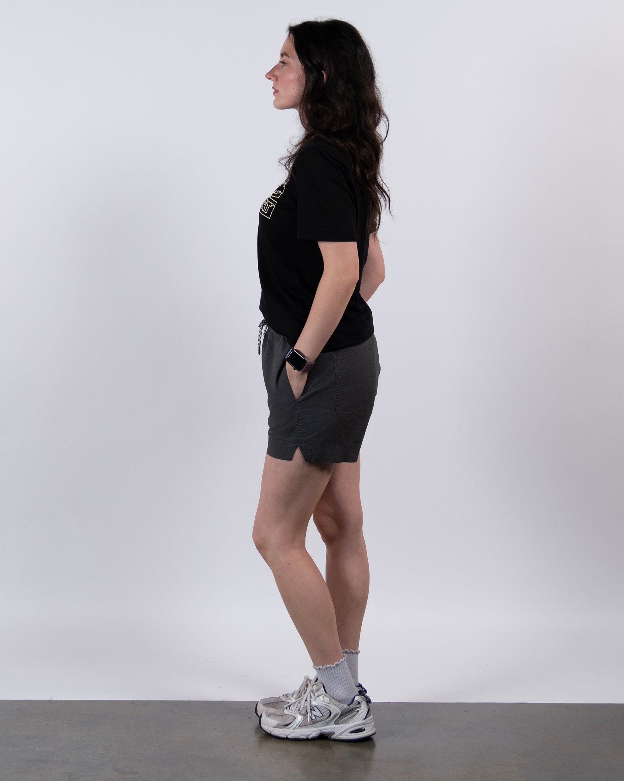 A woman with long dark hair stands in profile against a plain white background, wearing BEARDEDGOAT's Women's Canopy Short in gray stretch cotton ripstop, a black t-shirt, white socks, and gray sneakers.