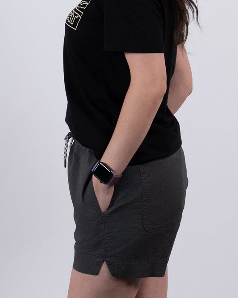 A woman with long dark hair stands in profile against a plain white background, wearing BEARDEDGOAT's Women's Canopy Short in gray stretch cotton ripstop, a black t-shirt, white socks, and gray sneakers.