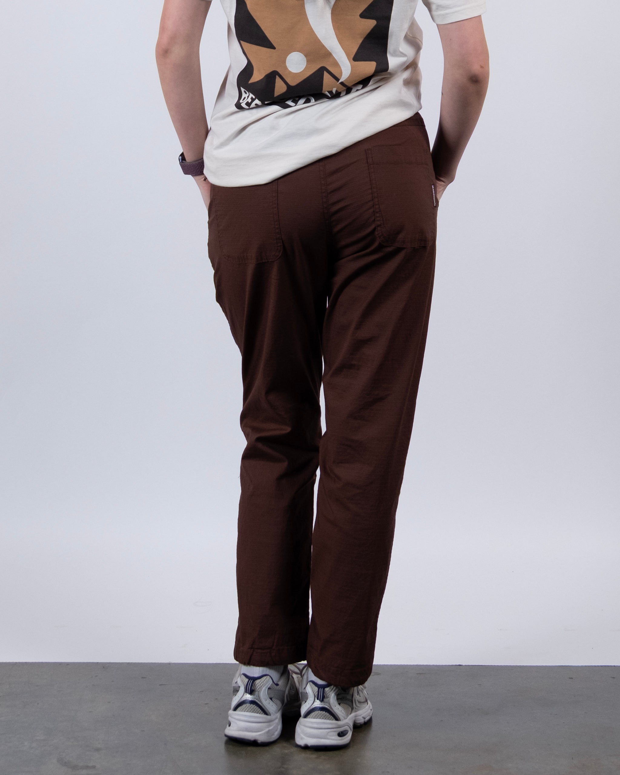 A person stands facing away, wearing a white graphic tee and BEARDEDGOAT Women's Canopy™ Pant in brown stretch cotton ripstop with adjustable ankle drawcords. White sneakers complete the look against a light-colored background.
