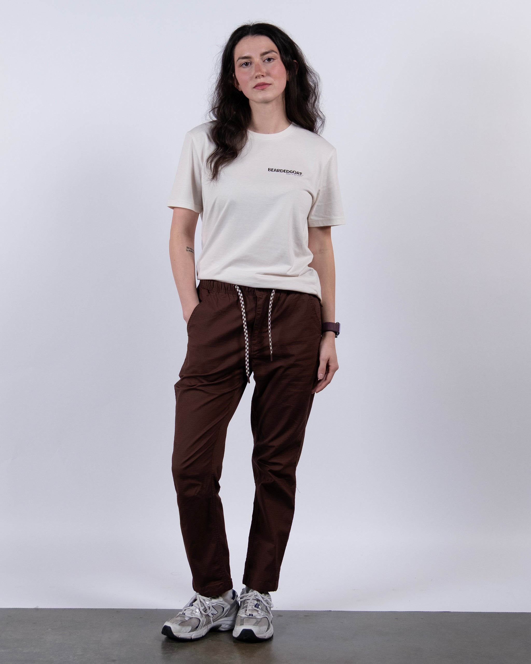 A woman with long dark hair stands against a white background wearing a cream tee, gray sneakers, and BEARDEDGOAT Women's Canopy™ Pant in brown with adjustable ankle drawcords. She has one hand in her pocket and looks relaxed.