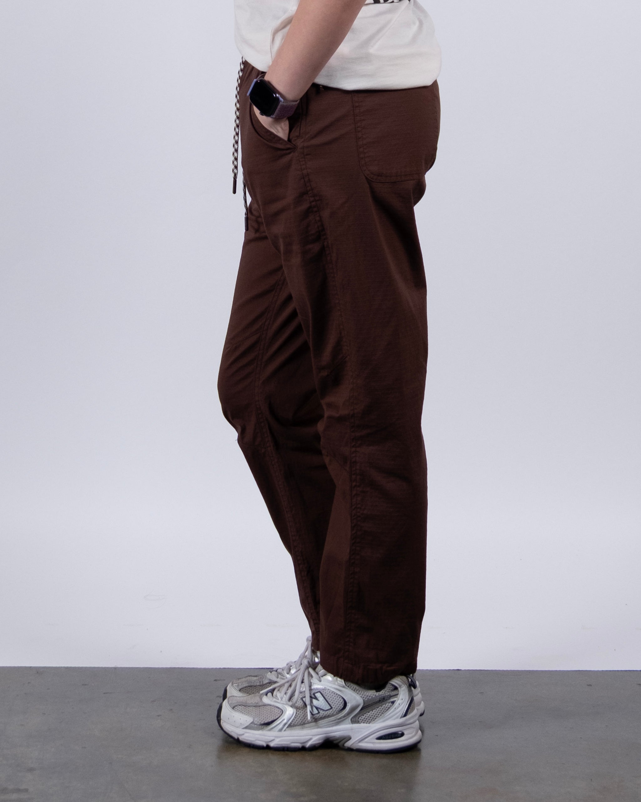 A person stands sideways with one hand in their pocket, wearing BEARDEDGOAT Women's Canopy™ Pant with adjustable ankle drawcords, a light shirt, a smartwatch, and gray athletic shoes against a plain light background.