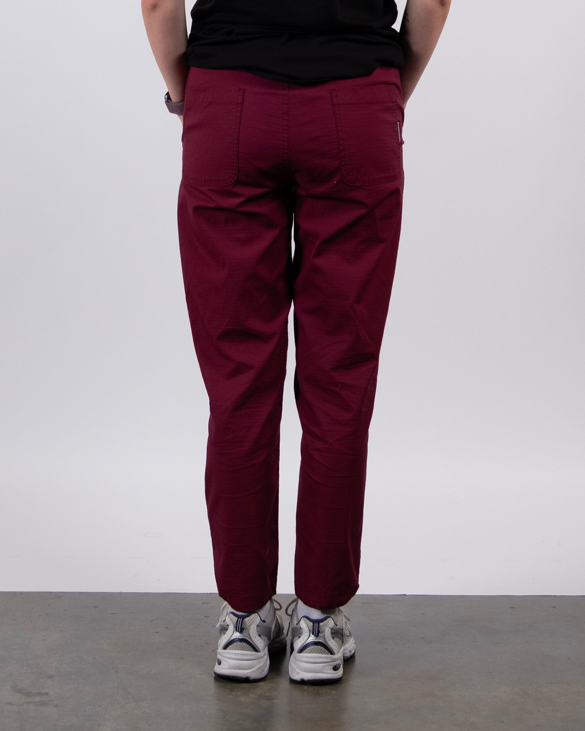 A person faces away from the camera wearing BEARDEDGOAT Women's Canopy™ Pant in burgundy with adjustable ankle drawcords, paired with a black shirt and white sneakers, standing against a plain light background and concrete floor.