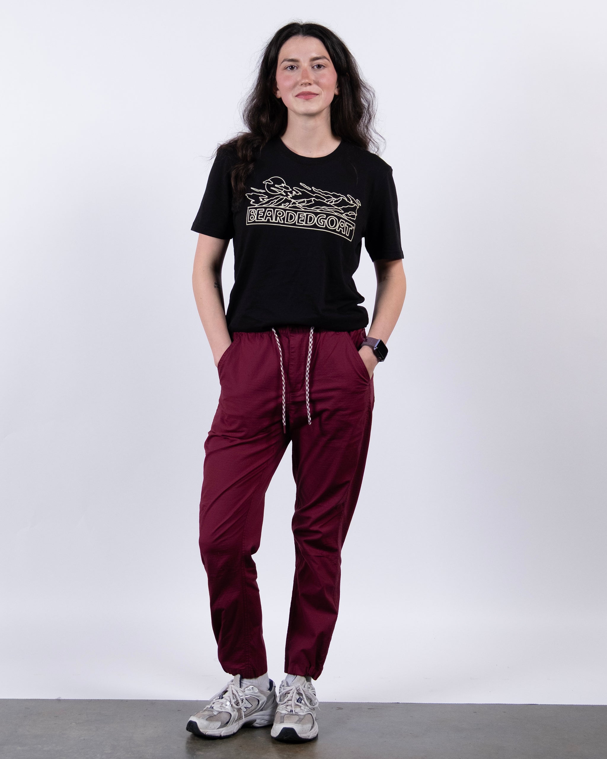 A person stands against a white background wearing a black BEARDED GOONS tee, BEARDEDGOAT Women's Canopy™ Pant in maroon with adjustable ankle drawcords, gray sneakers, and a smartwatch, hands in pockets.