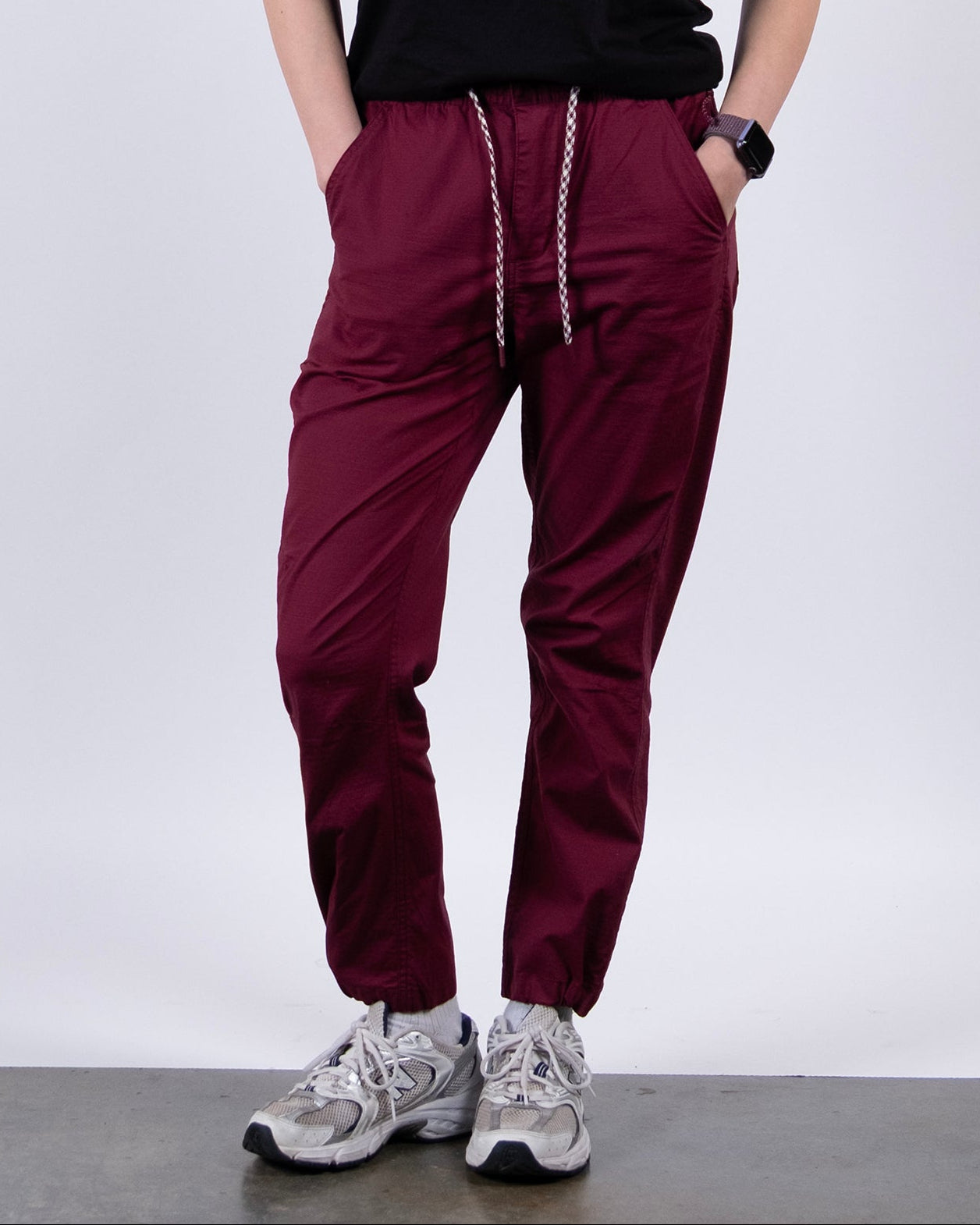 A person wearing BEARDEDGOAT Women's Canopy™ Pant in burgundy, featuring adjustable ankle drawcords, pairs it with a black top, gray sneakers, and a smartwatch while standing with hands in pockets against a plain white background.
