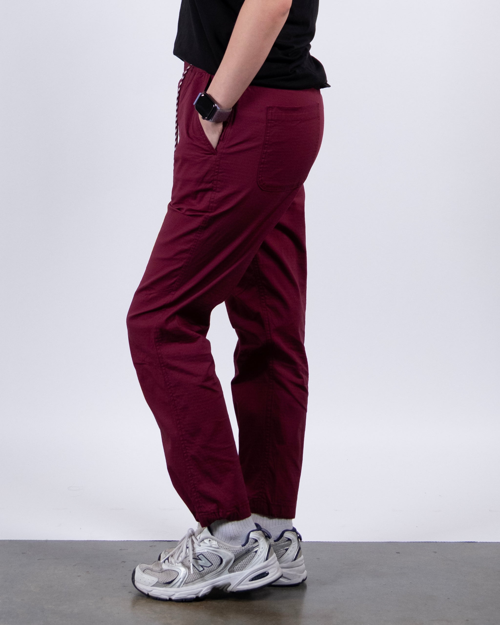 A person stands sideways in a black shirt, BEARDEDGOAT Women's Canopy™ Pant with adjustable ankle drawcords (maroon), white sneakers, and a smartwatch on their left wrist against a plain light background.